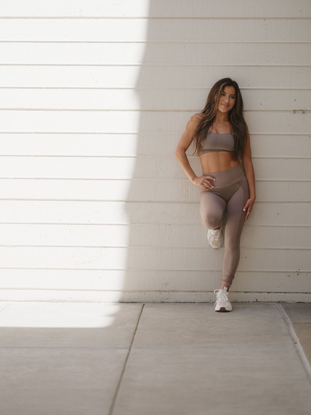 Athletic woman in taupe workout set poses against white wall with dramatic lighting.