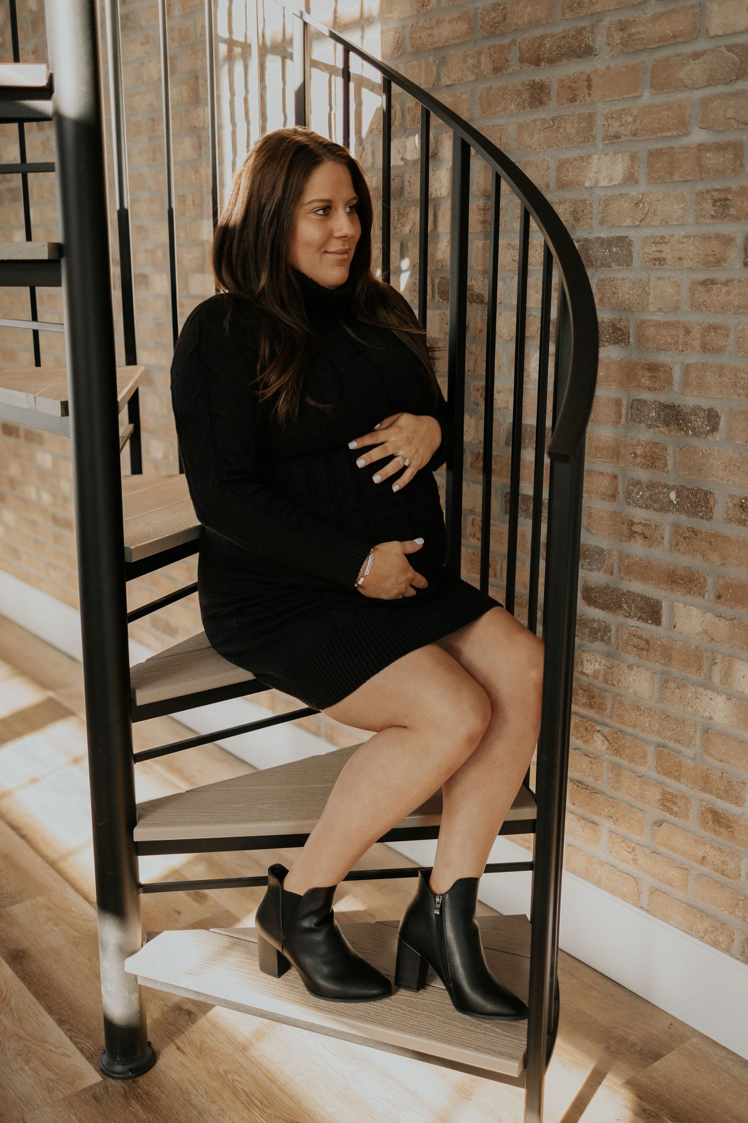 Person in a black sweater dress and ankle boots poses elegantly on spiral staircase against exposed brick wall at Minnesota studio.