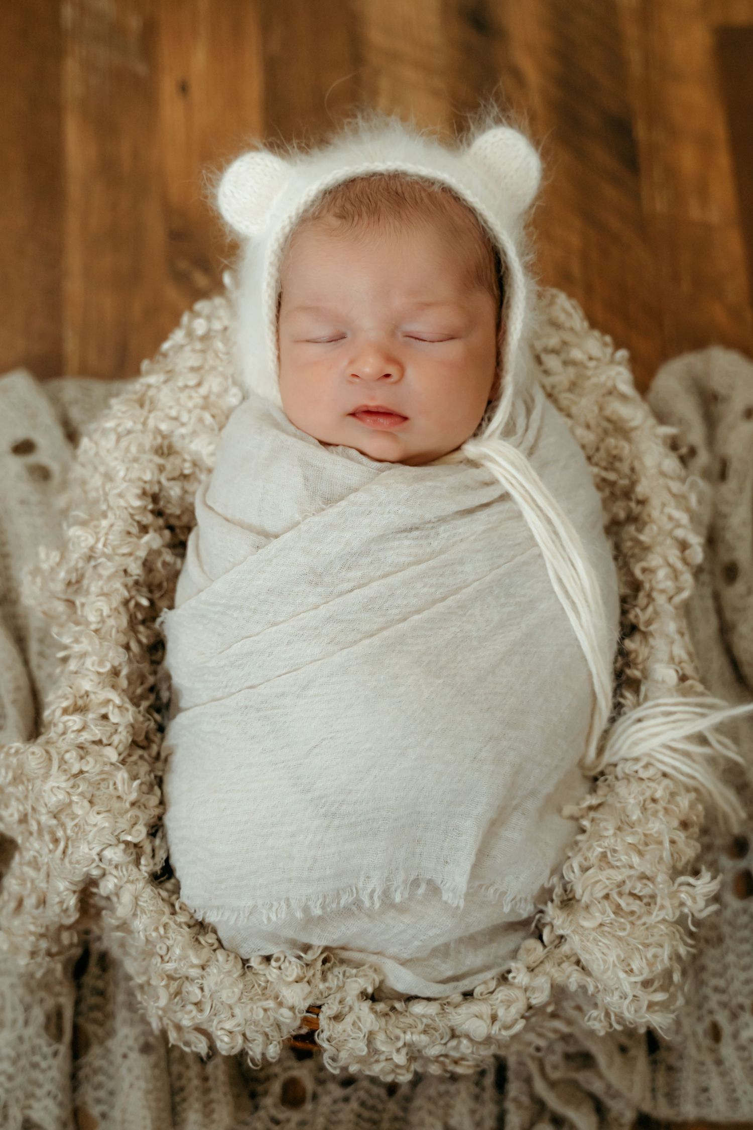 Newborn in white bear bonnet sleeps peacefully swaddled on textured blanket.