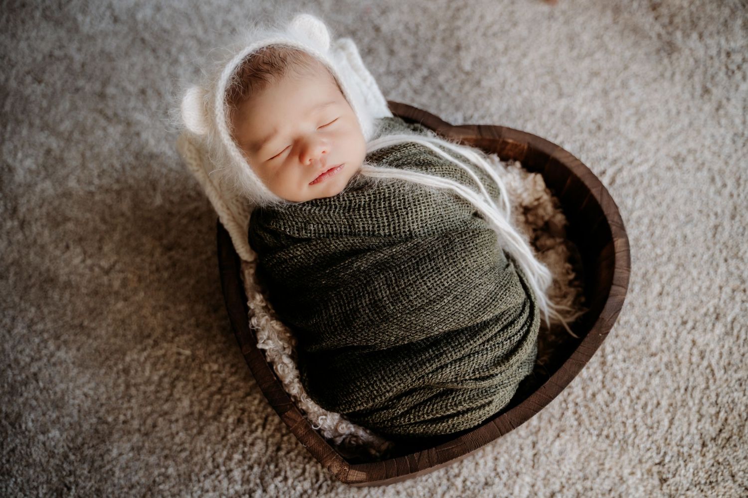 Sleeping newborn wrapped in gray knit swaddle rests in wooden bowl.