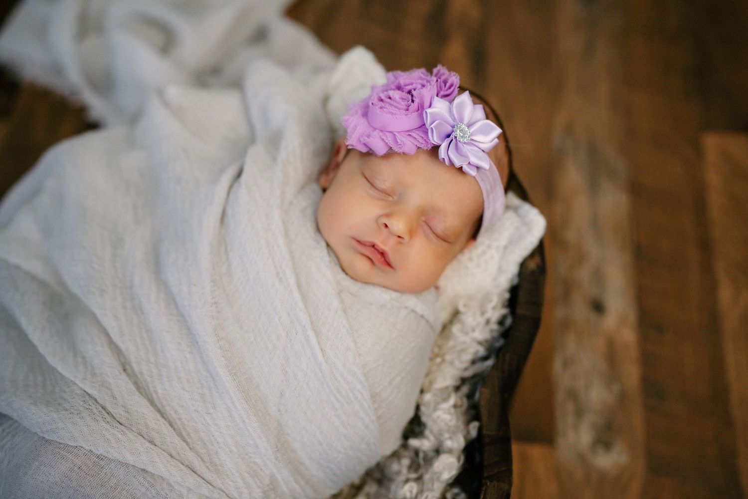 Sleeping newborn with pink floral headband wrapped in white swaddle.