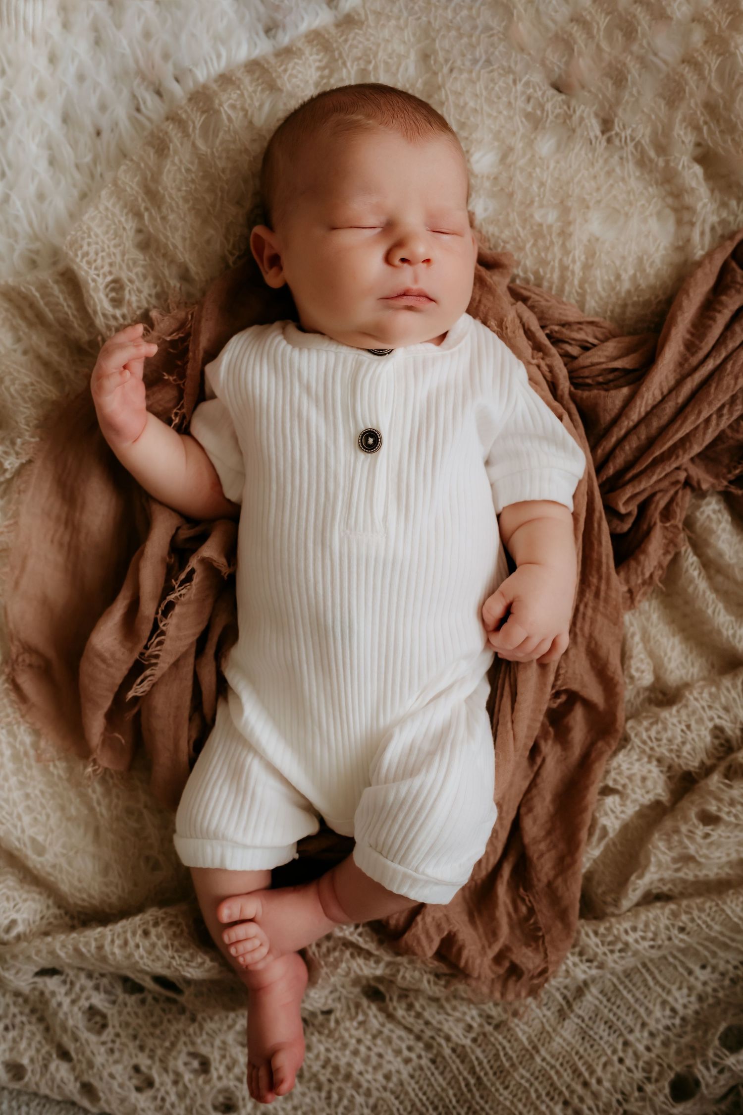 Sleeping baby wearing white ribbed romper rests on brown blanket.