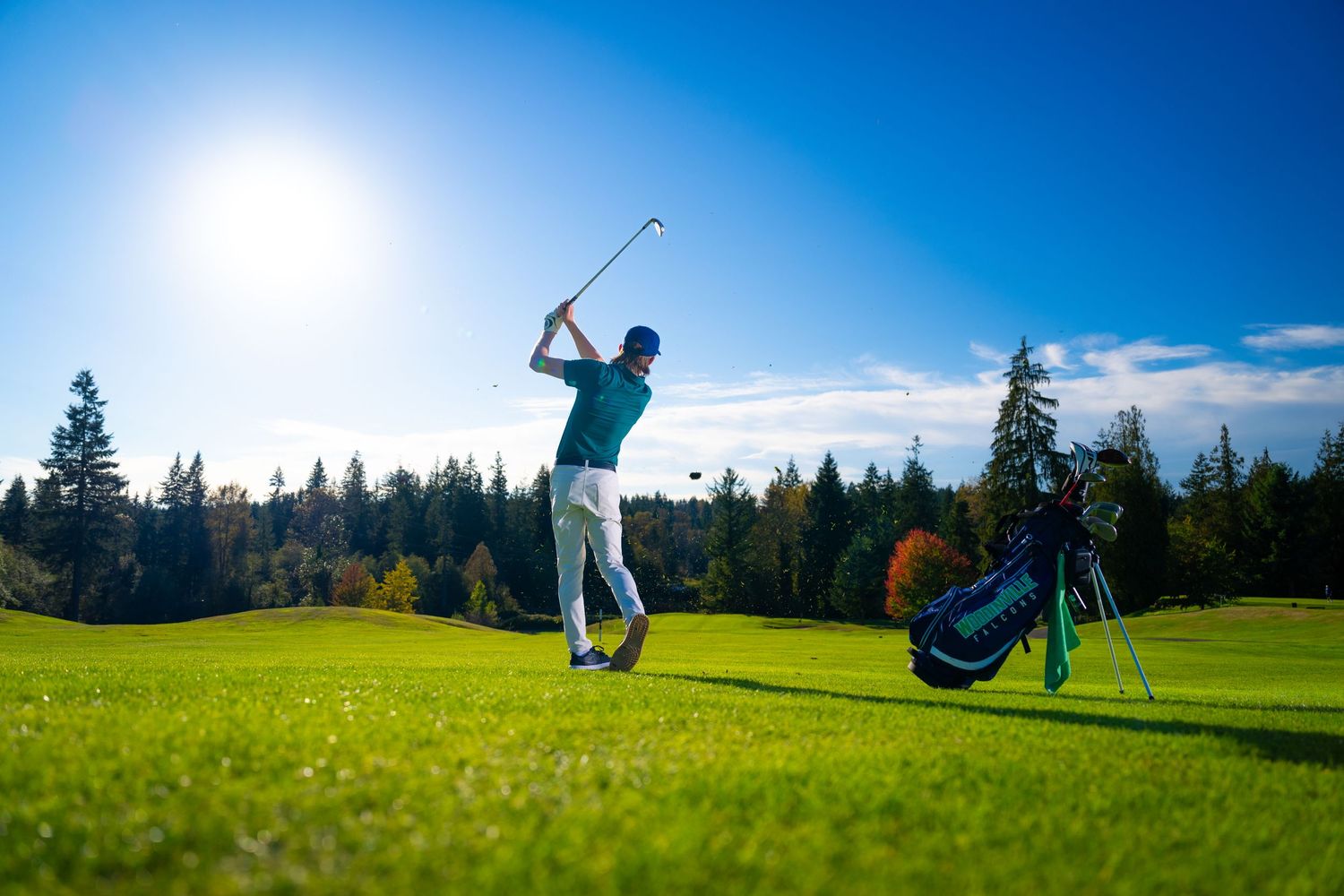 A golfer takes a swing on a lush green course with golf bag and evergreen trees in the background on a sunny day.