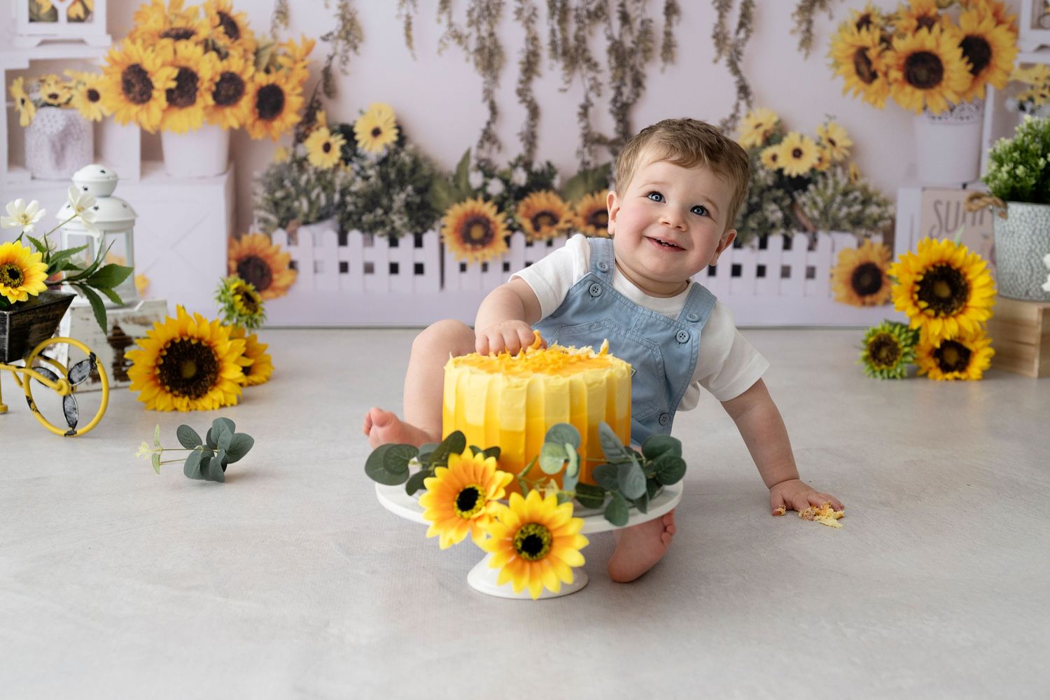 Cake Smash Photography, a toddler wearing blue romper, sitting behind a yellow birthday cake surrounded by bright sunflowers.