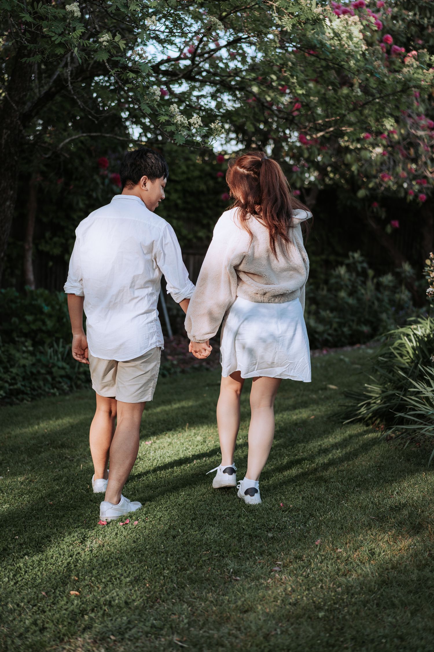 A couple in white outfits walks hand in hand through a lush garden path.