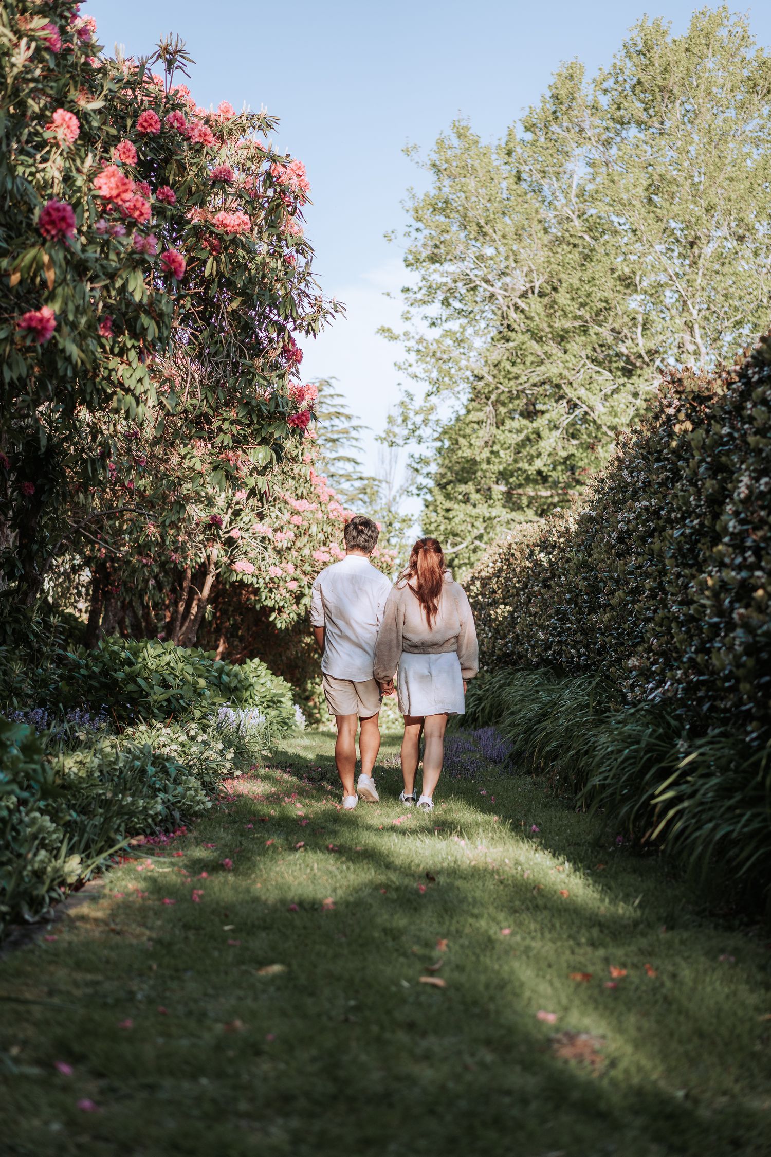 A couple walks down a garden path lined with flowering trees and lush greenery.