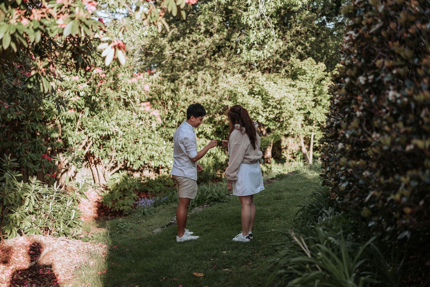 Two people share a special moment in a sunlit garden surrounded by lush greenery and flowers.