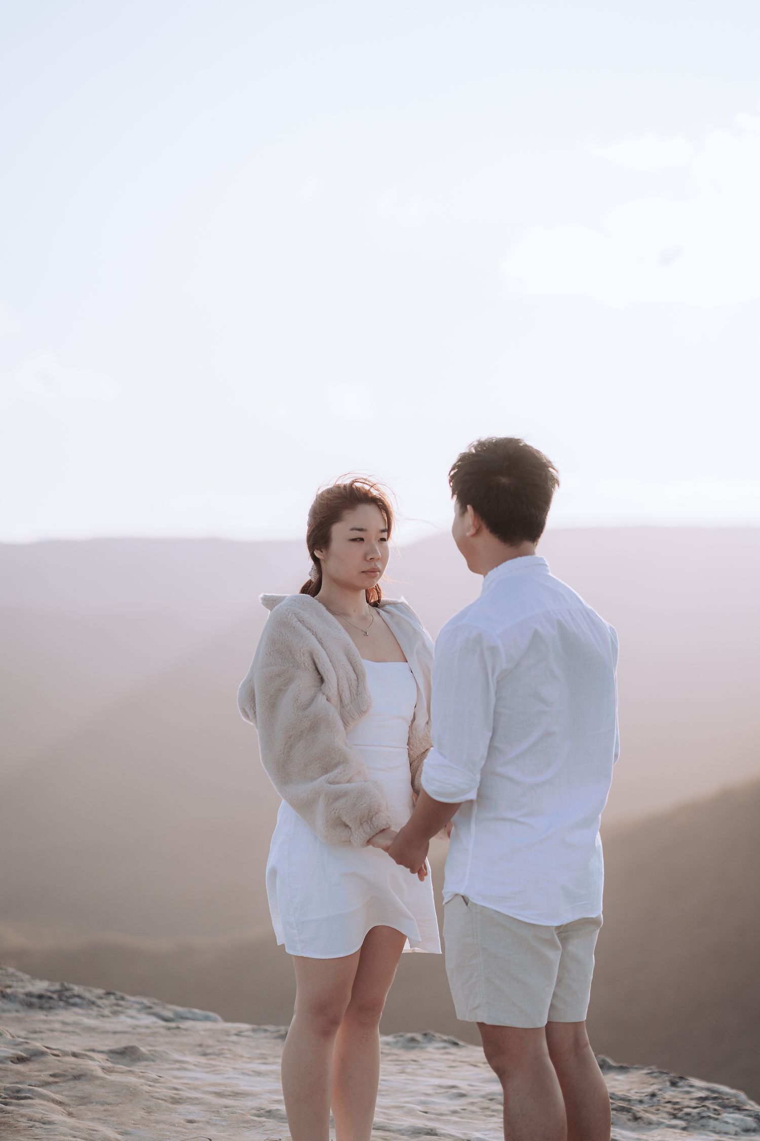 Two people in white outfits share a romantic moment on a cliff edge during sunset.