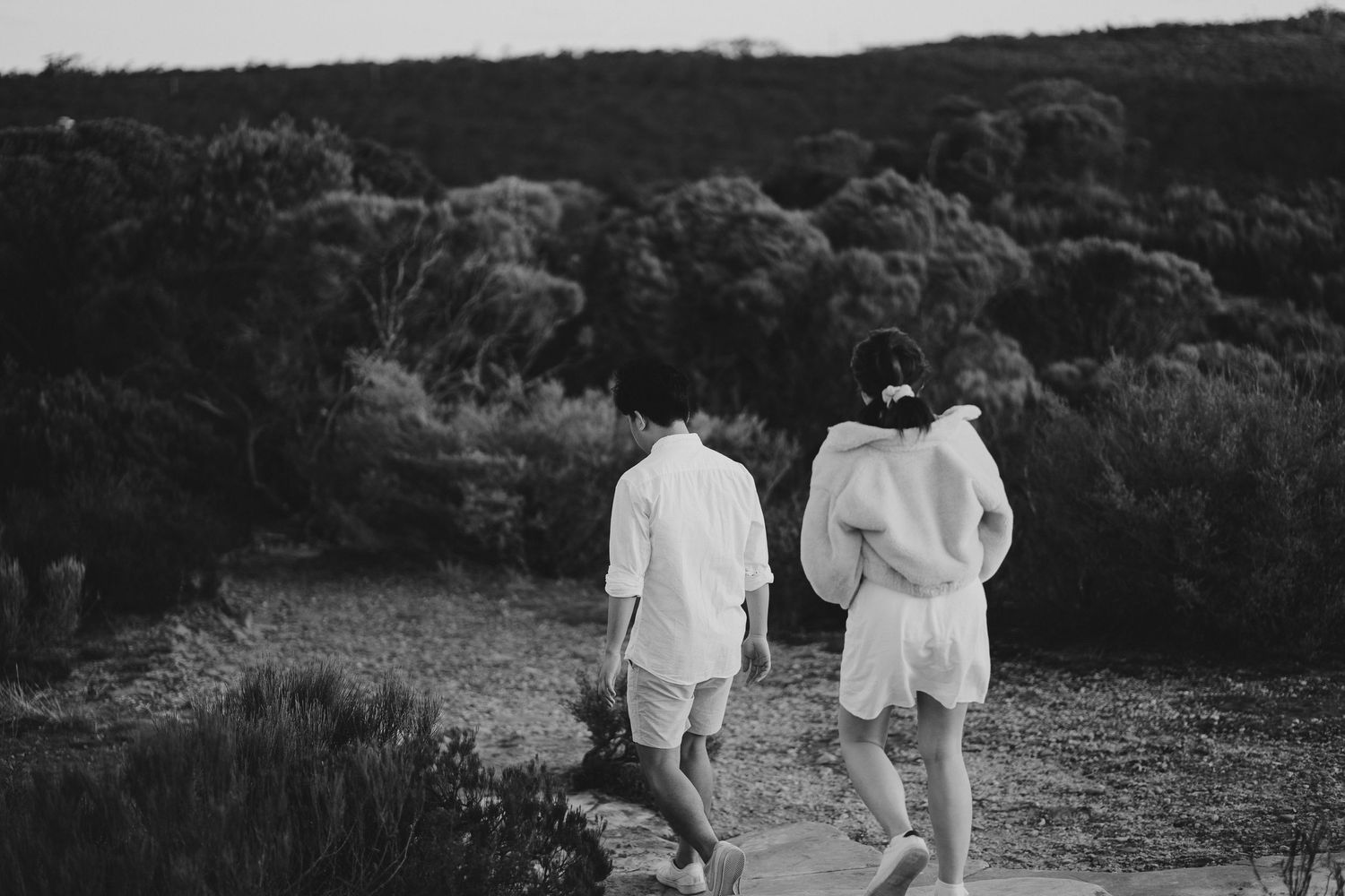 A couple in white outfits walking along a scenic hiking trail surrounded by lush greenery.