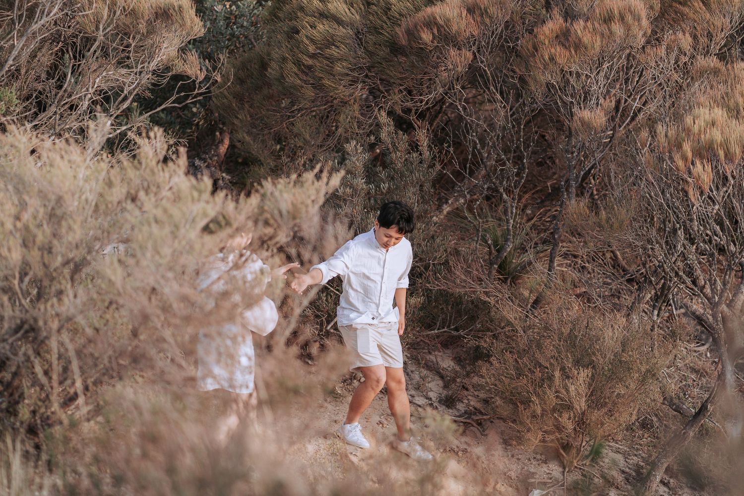 Two figures in white clothing exploring a rugged mountain trail through dried brush.