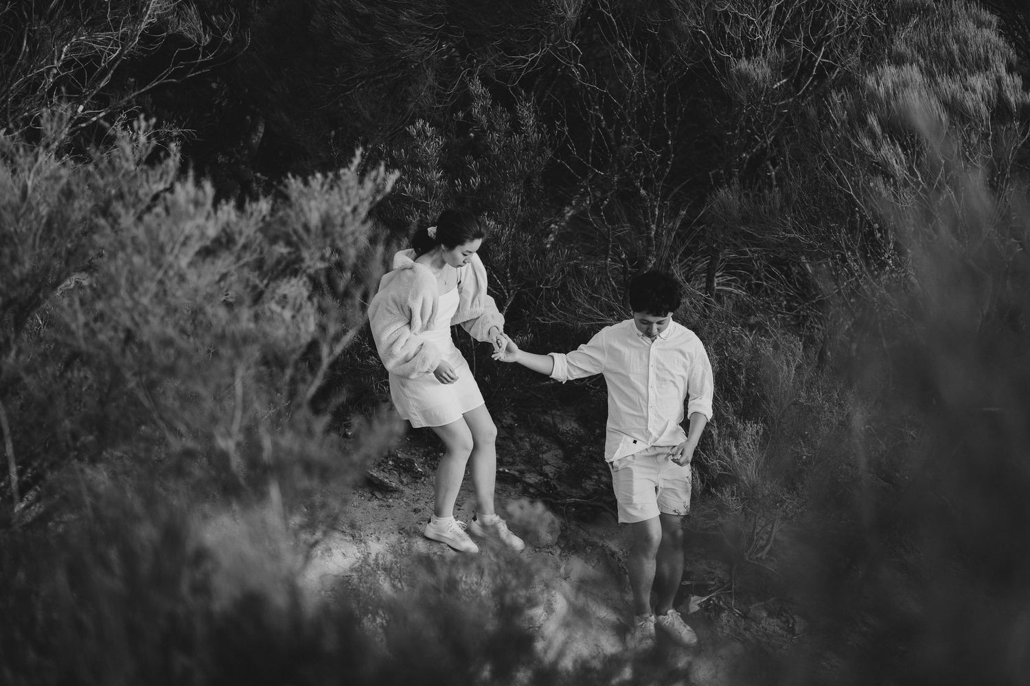 Couple in white attire helping each other traverse rocky terrain on a hiking adventure.