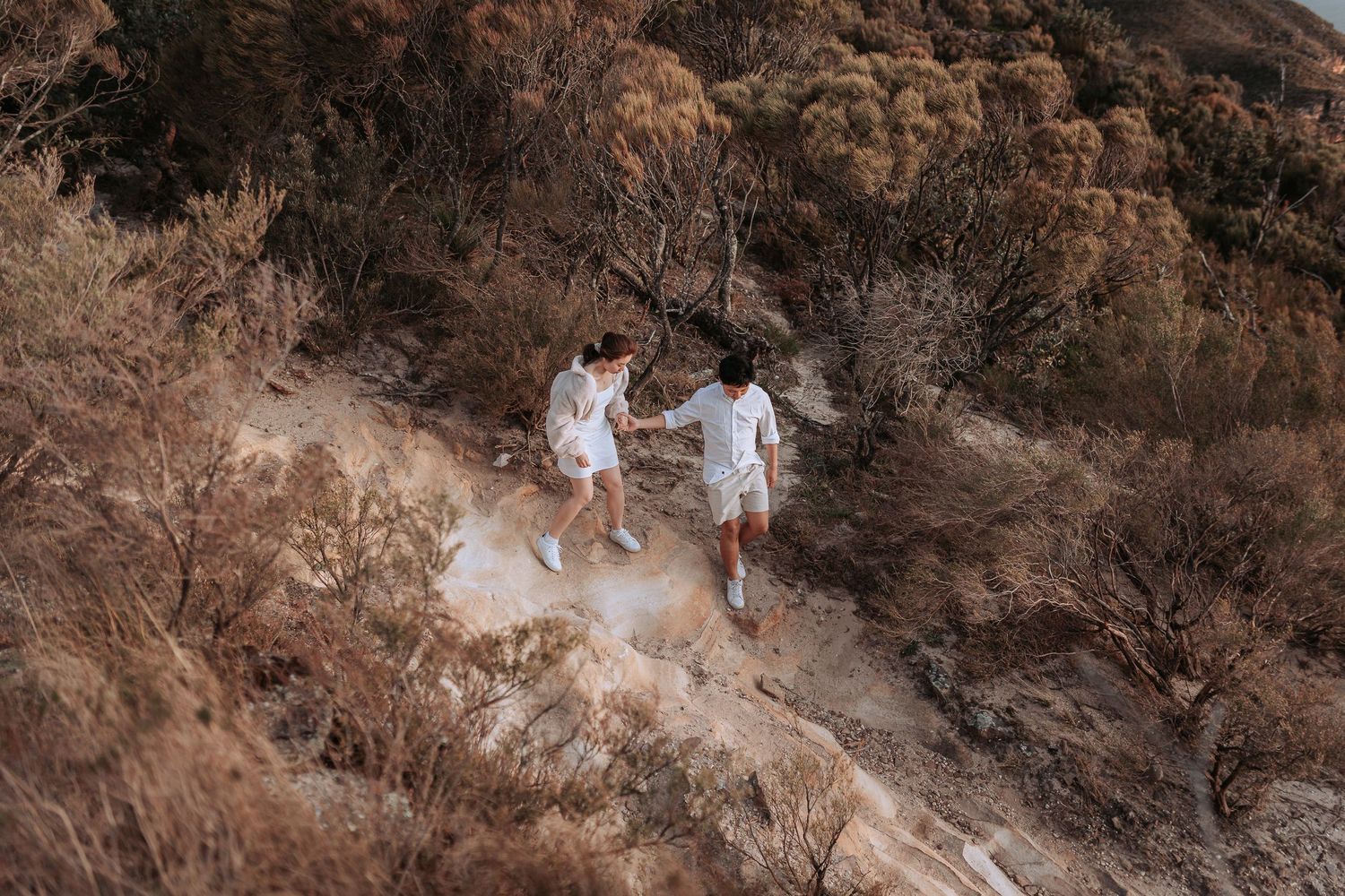 A couple in white clothing walking together on a dusty mountain path.