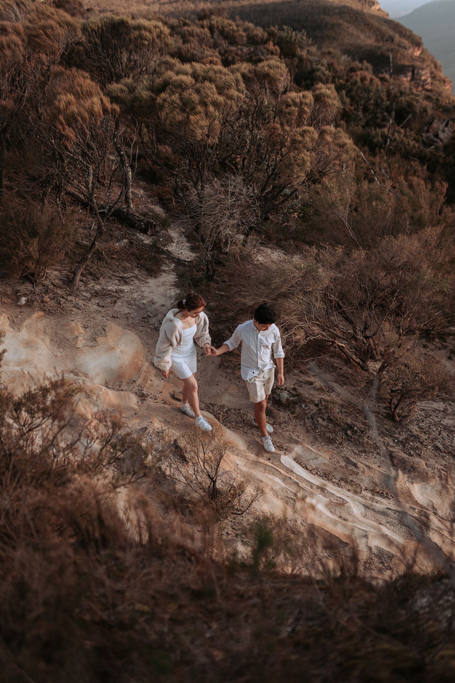 A couple holding hands while hiking down a scenic mountain trail in matching white outfits.