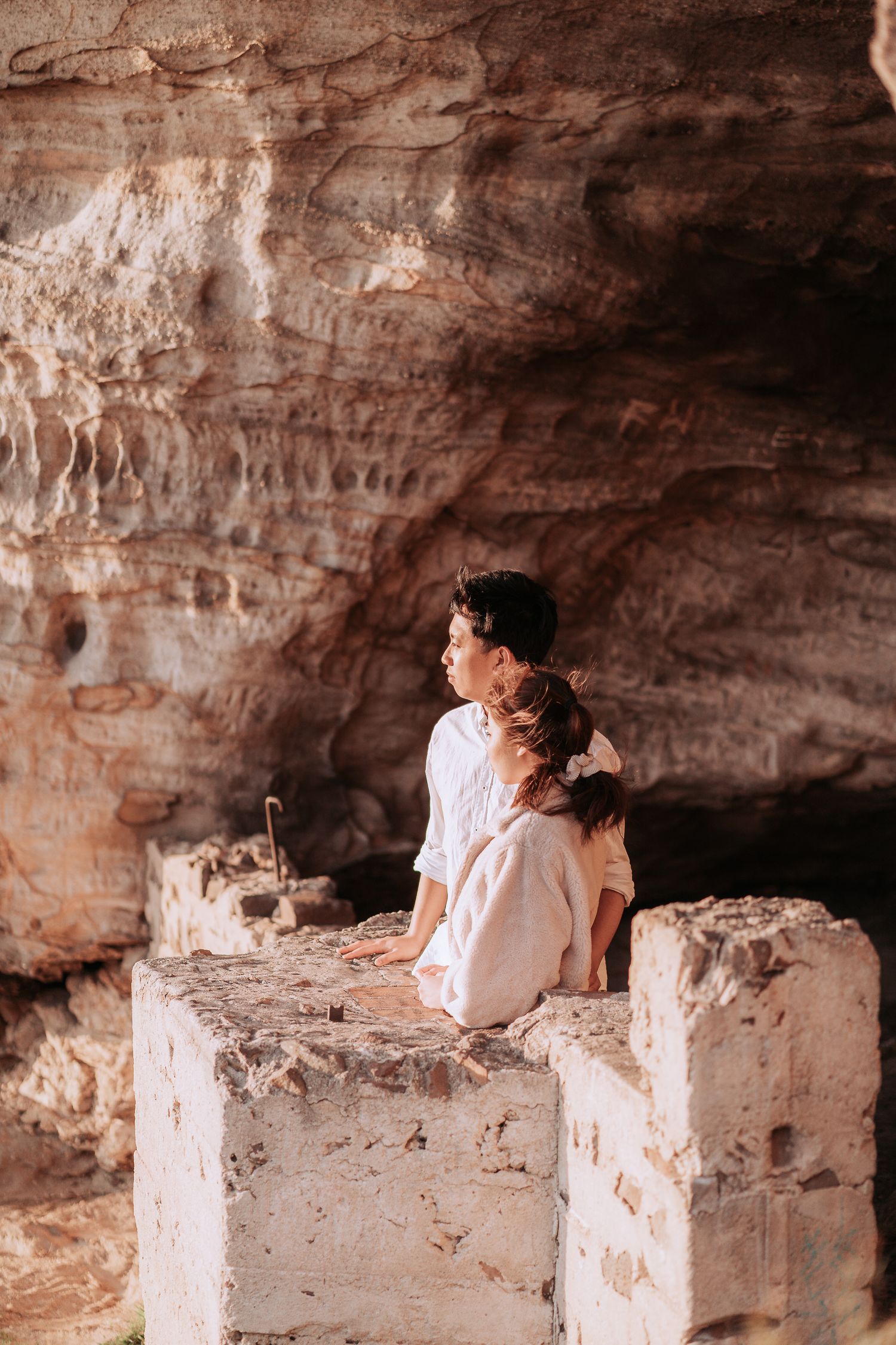 Two people sitting together on a stone ledge inside a dramatic cave formation.