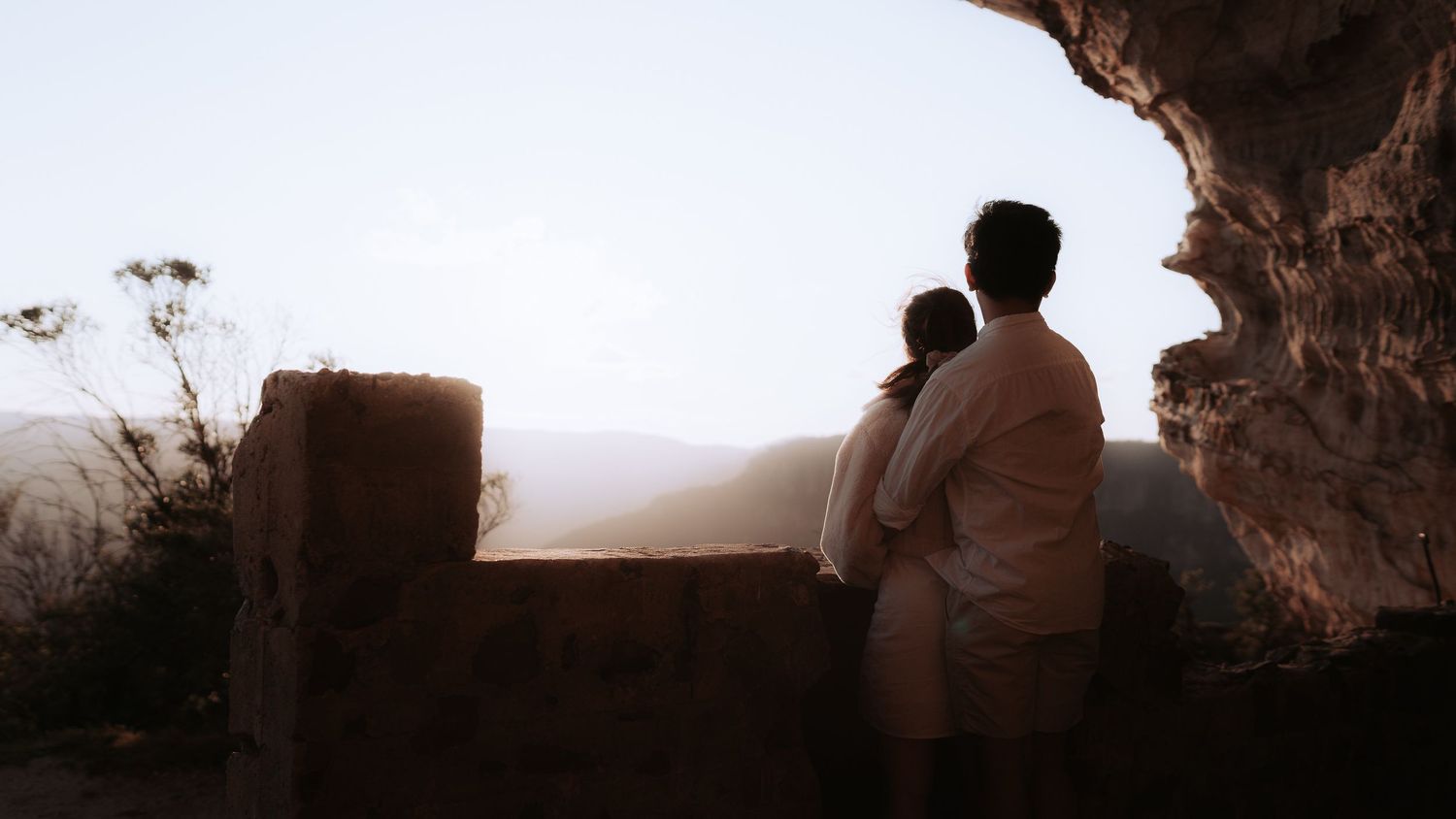 Two figures embracing while looking out over a misty valley from a cave opening.