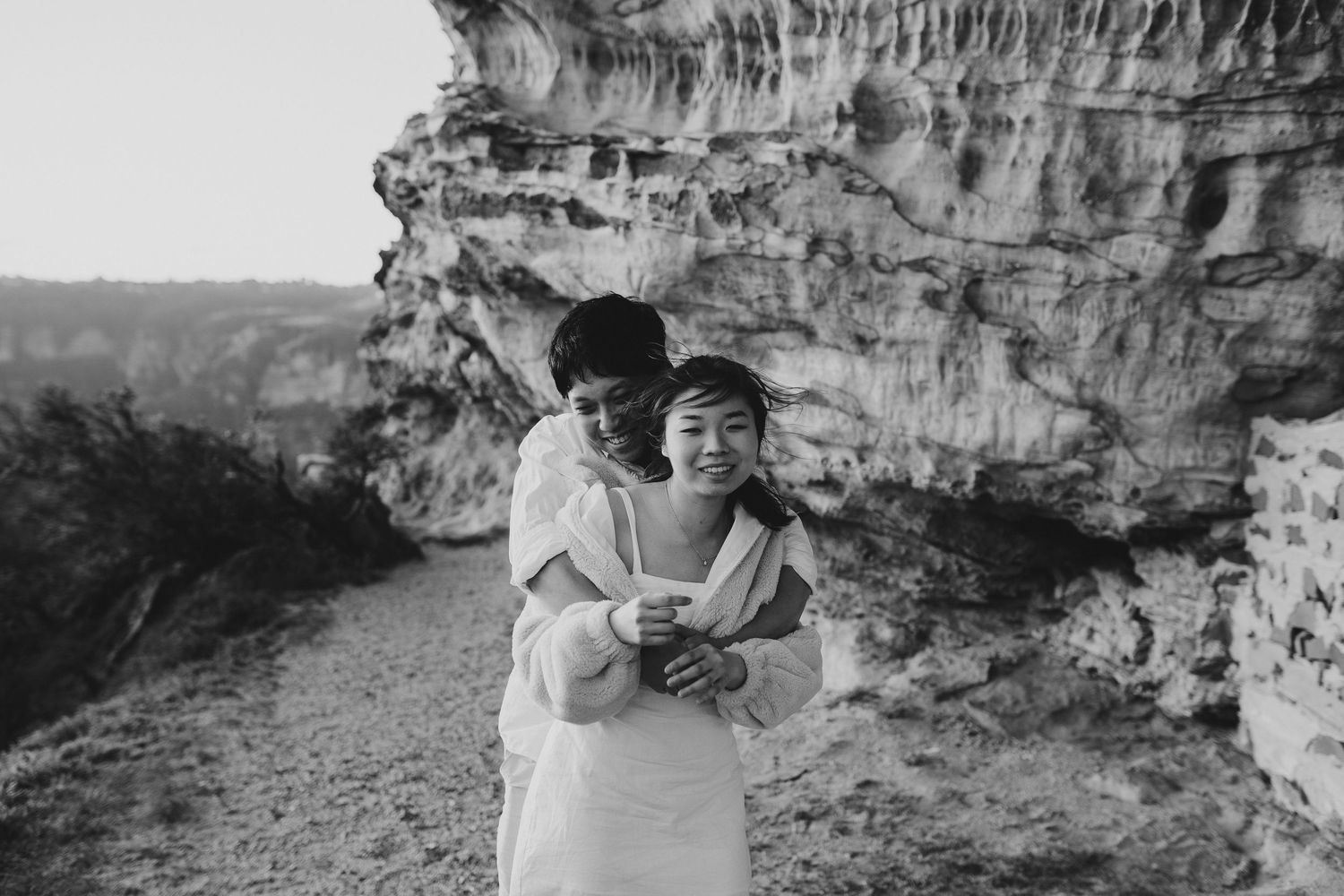 A black and white photo of a couple embracing on a rocky cliff path.
