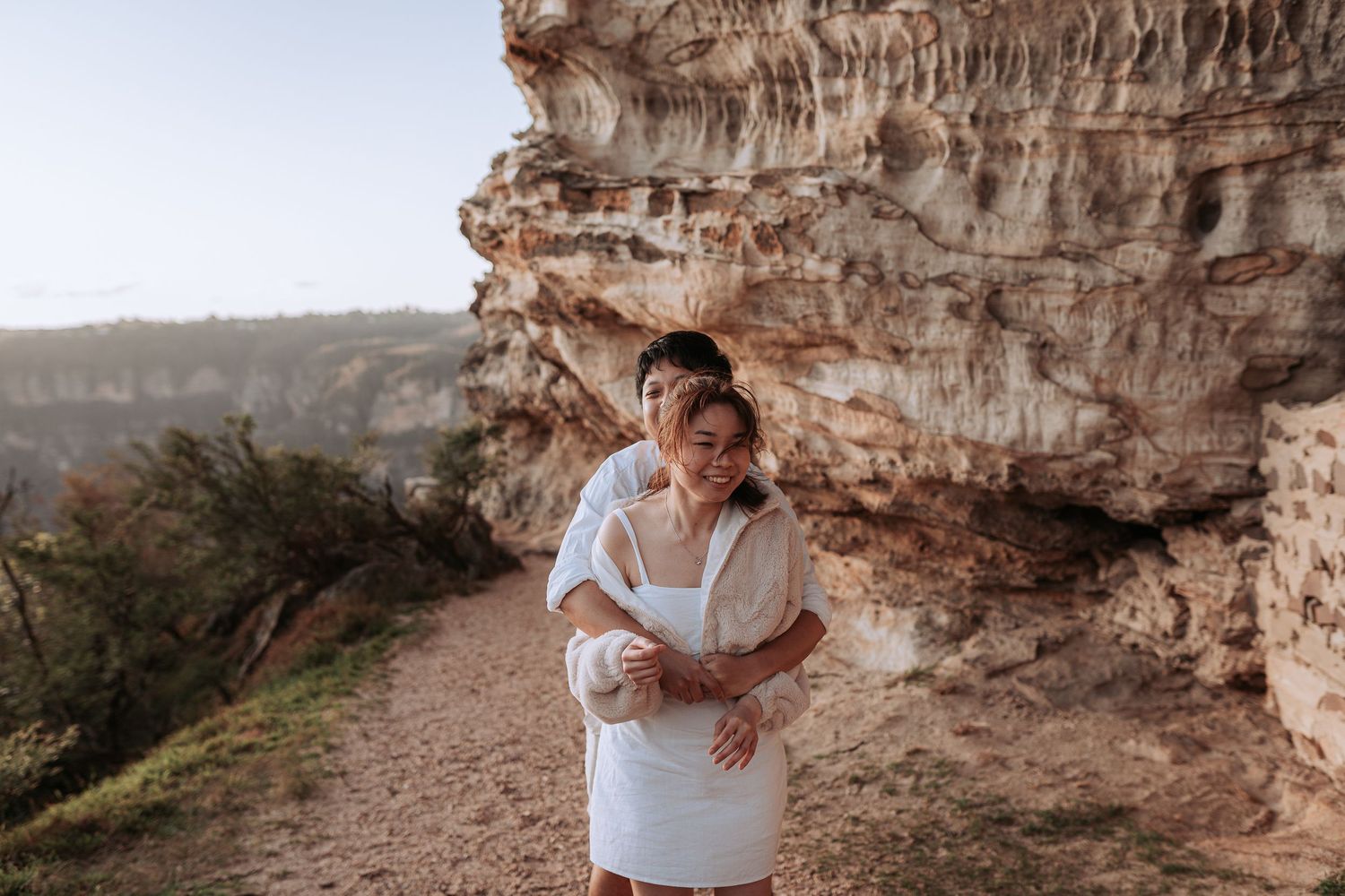 A couple in white outfits embracing against a dramatic rocky cliff backdrop in warm sunset light.