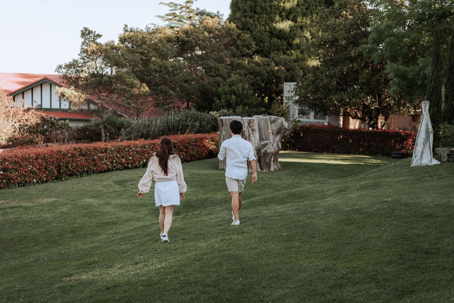 Two people walk together across a landscaped garden with trees and hedges in the background.