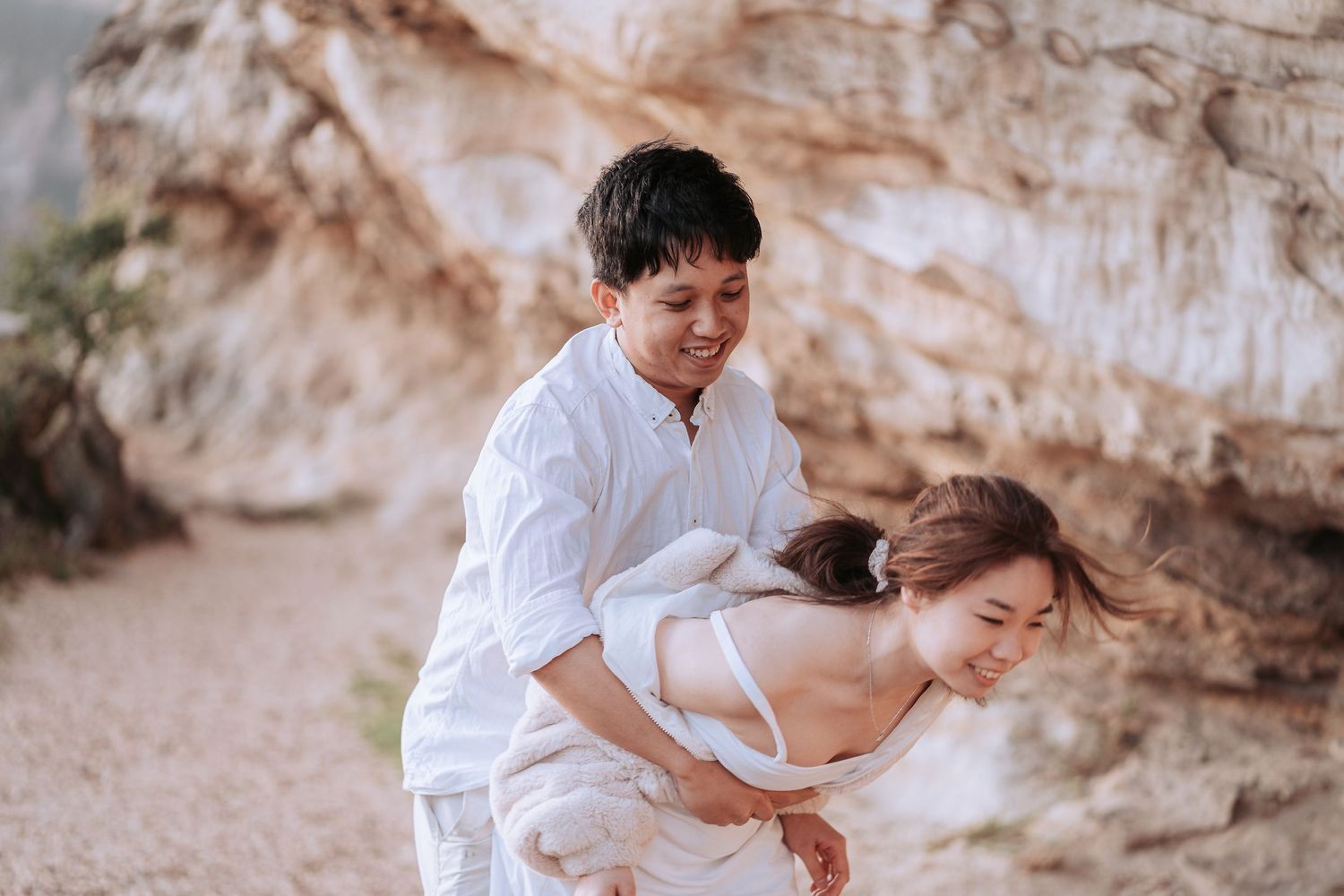 A romantic candid moment captured between a couple in white clothing near rocky cliffs.