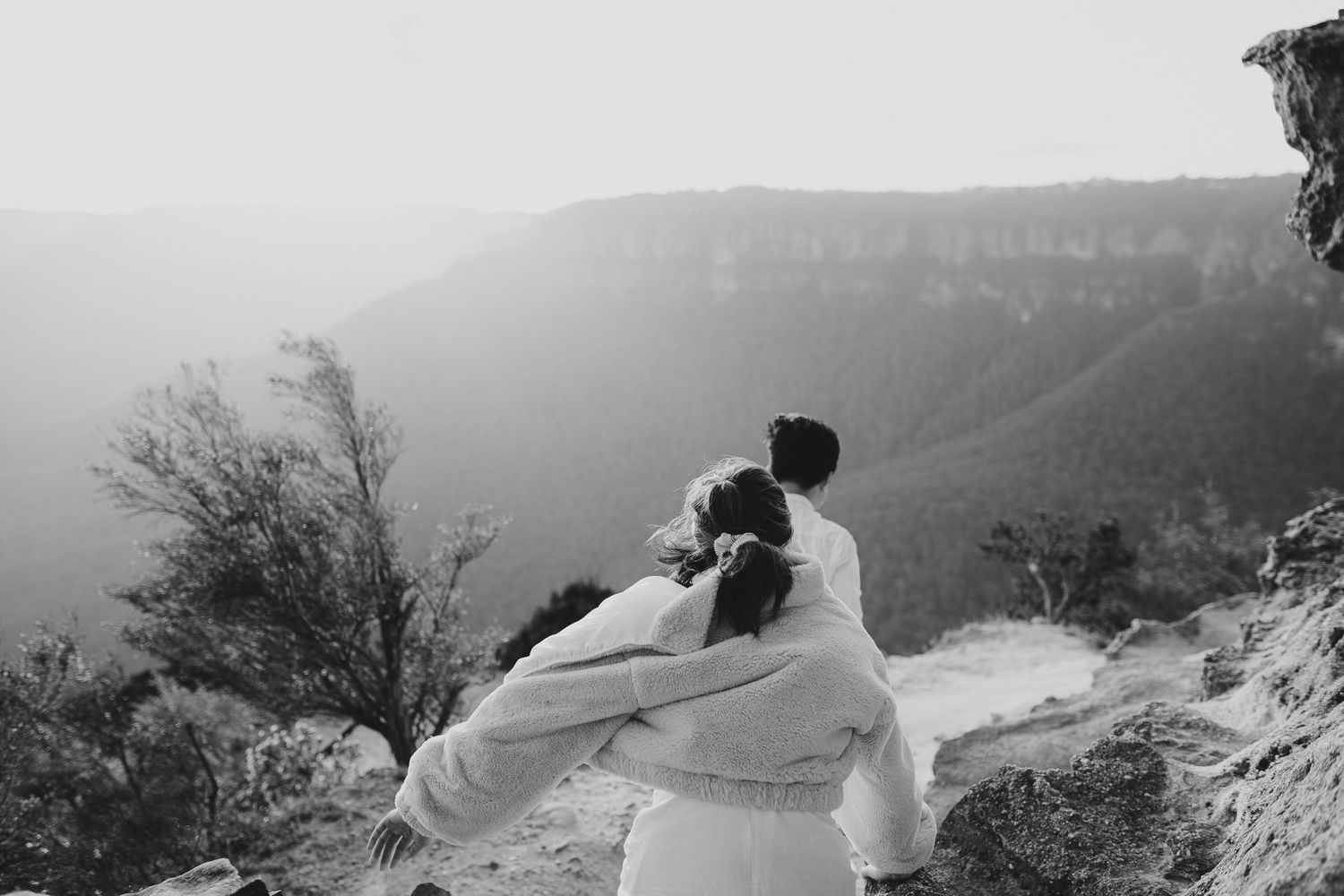 A black and white photo capturing a romantic moment on a mountain cliff edge.