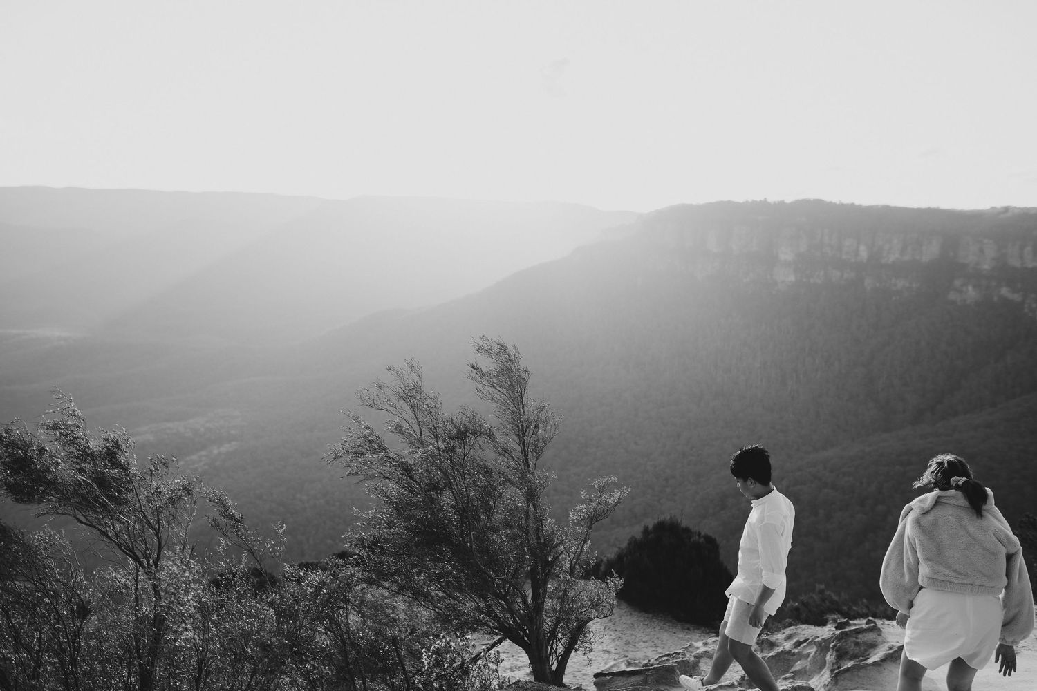 A black and white landscape photo of two people standing on a mountain viewpoint.