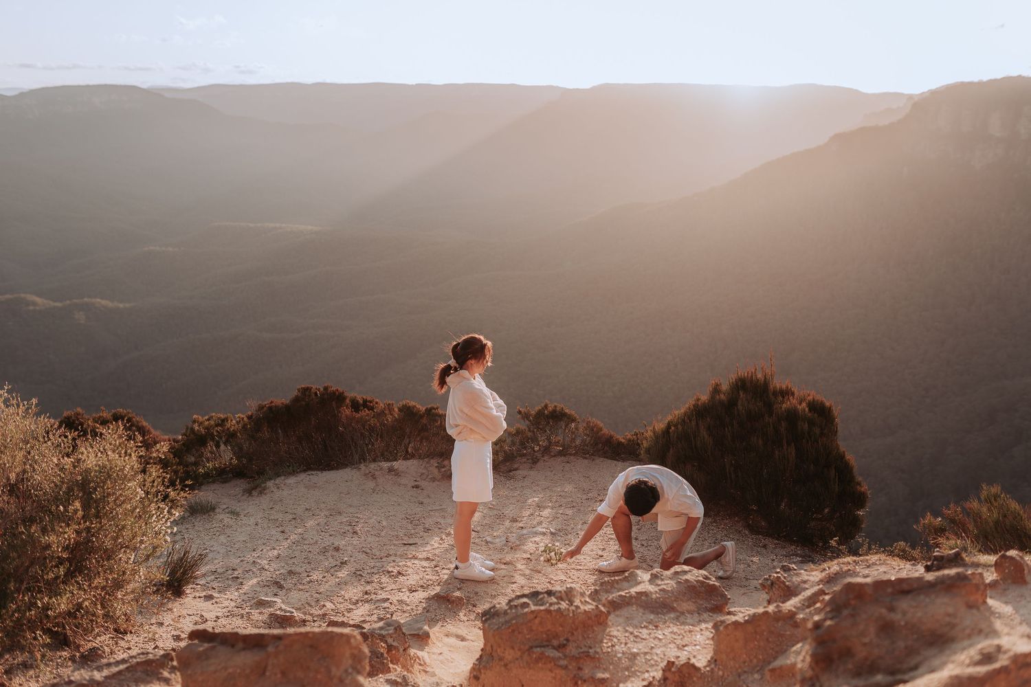 Two people in white clothing on a rocky mountain ledge during golden hour.
