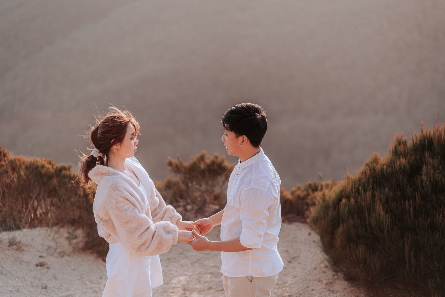 Couple in white clothing holding hands while standing on a cliff edge at sunset.