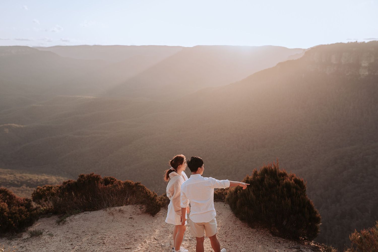 Couple pointing at the view while standing on a mountain cliff during golden hour.