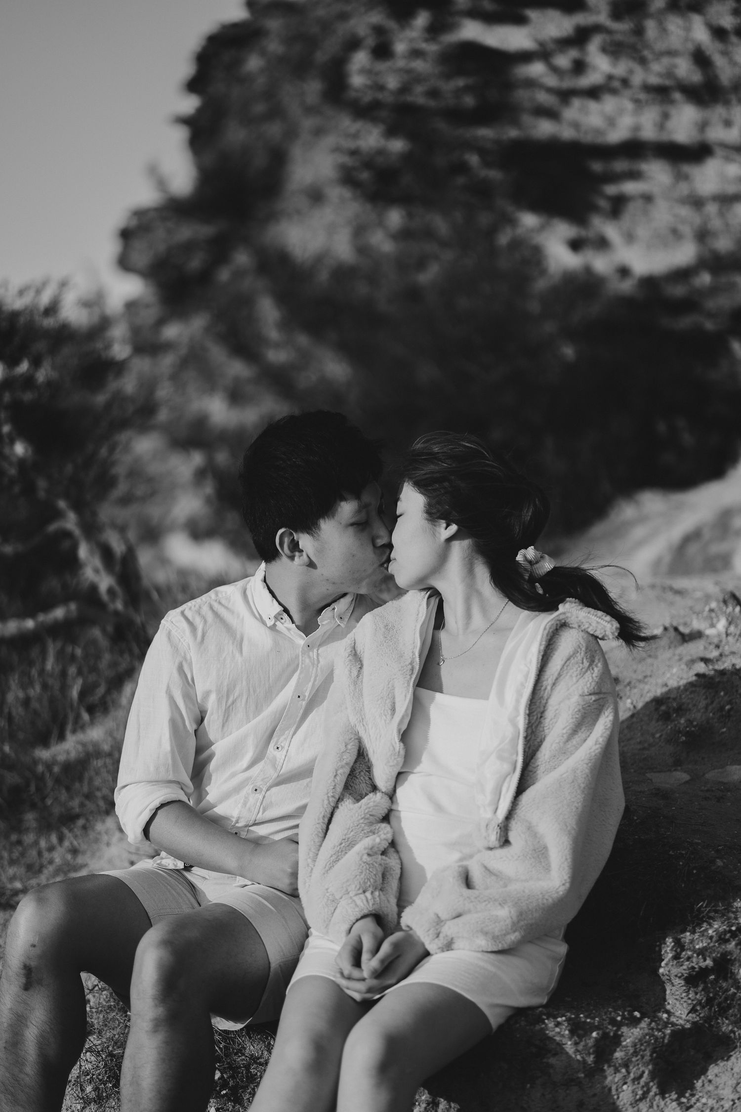 A romantic couple in white outfits sharing an intimate moment on rocky terrain at sunset.