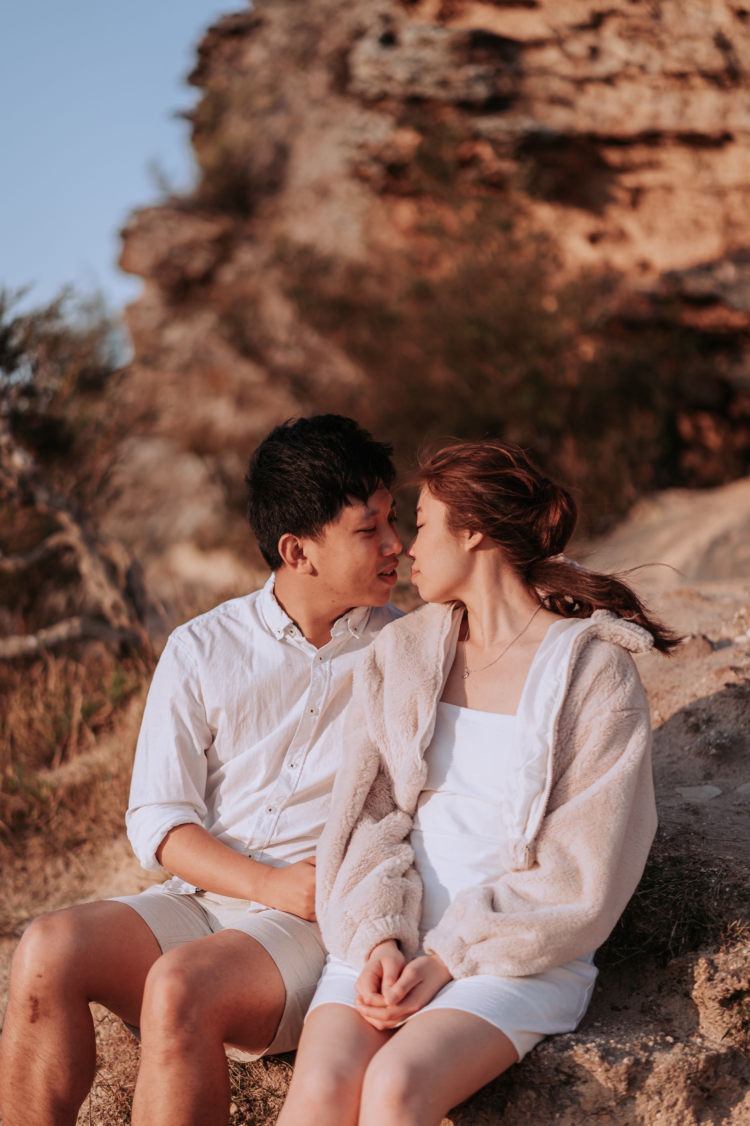 A couple dressed in white embraces while sitting on rocky cliffs during golden hour.