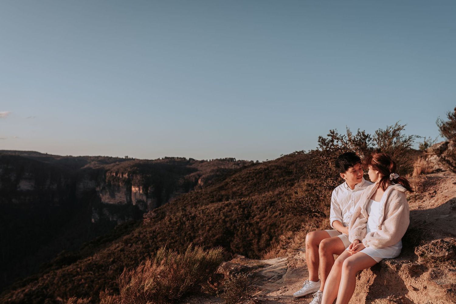 A couple in matching white outfits sits together on a scenic mountain overlook.