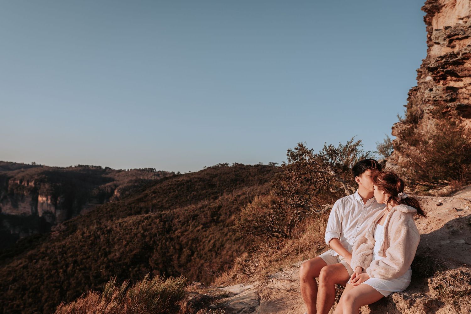 A couple in light-colored clothing sits together on a mountainside during sunset.
