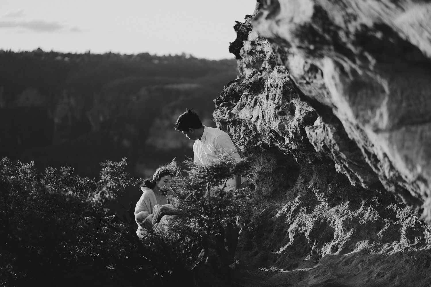 A black and white photo of two people sitting on a rocky cliff edge overlooking a valley.