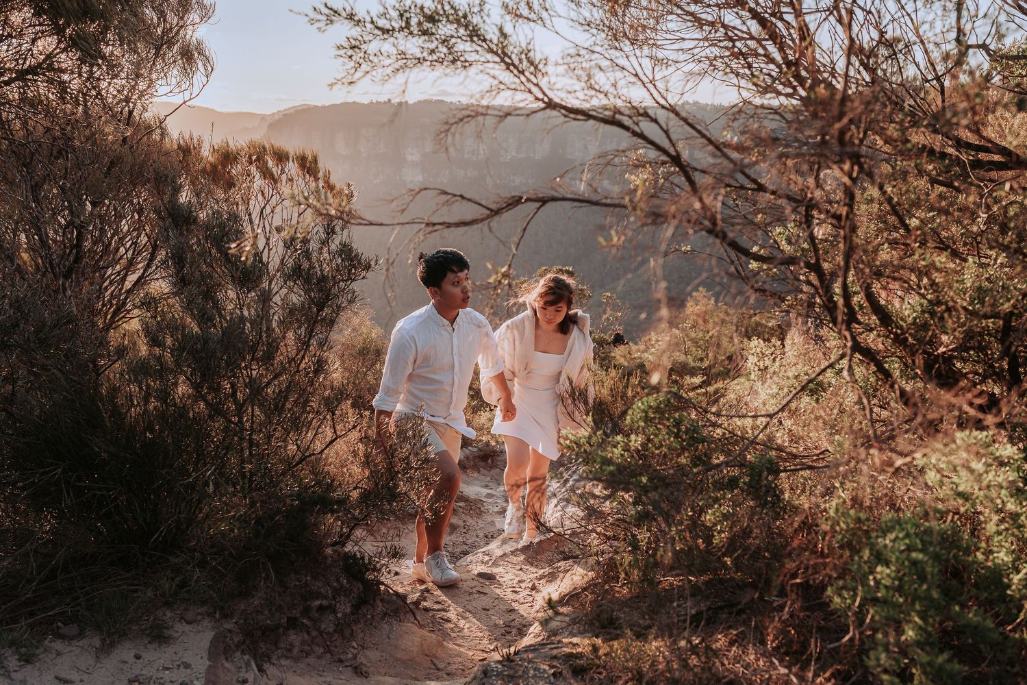 A couple in white outfits hiking together on a rocky mountain trail during golden hour.