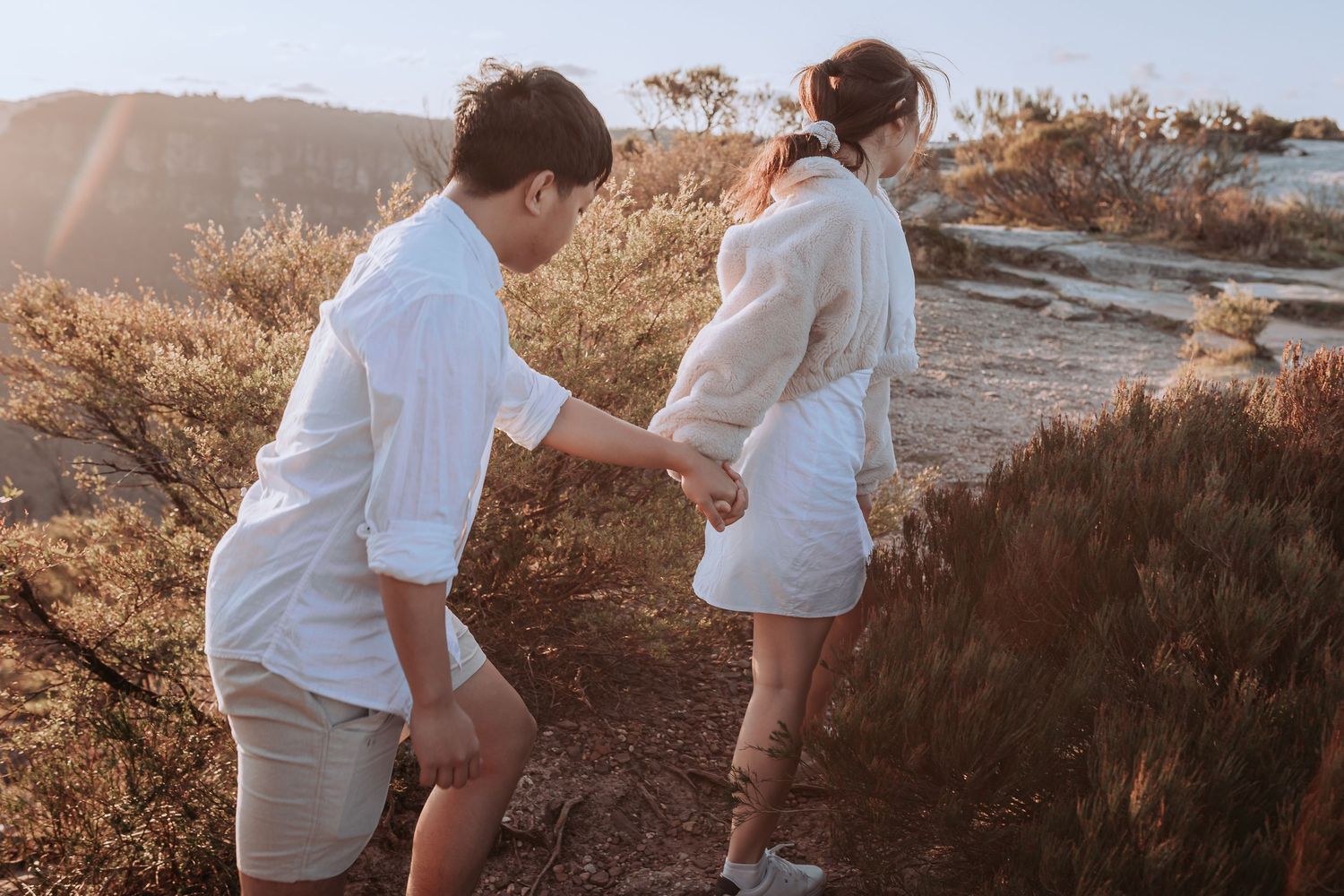 A couple in matching white outfits walks along a scenic mountain trail during sunset.