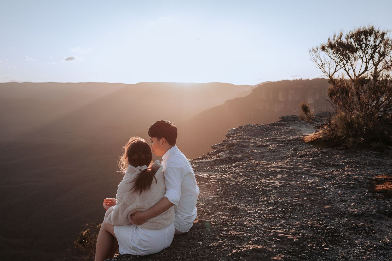 A romantic couple sits together on a cliff edge watching the sunset over mountain ranges.