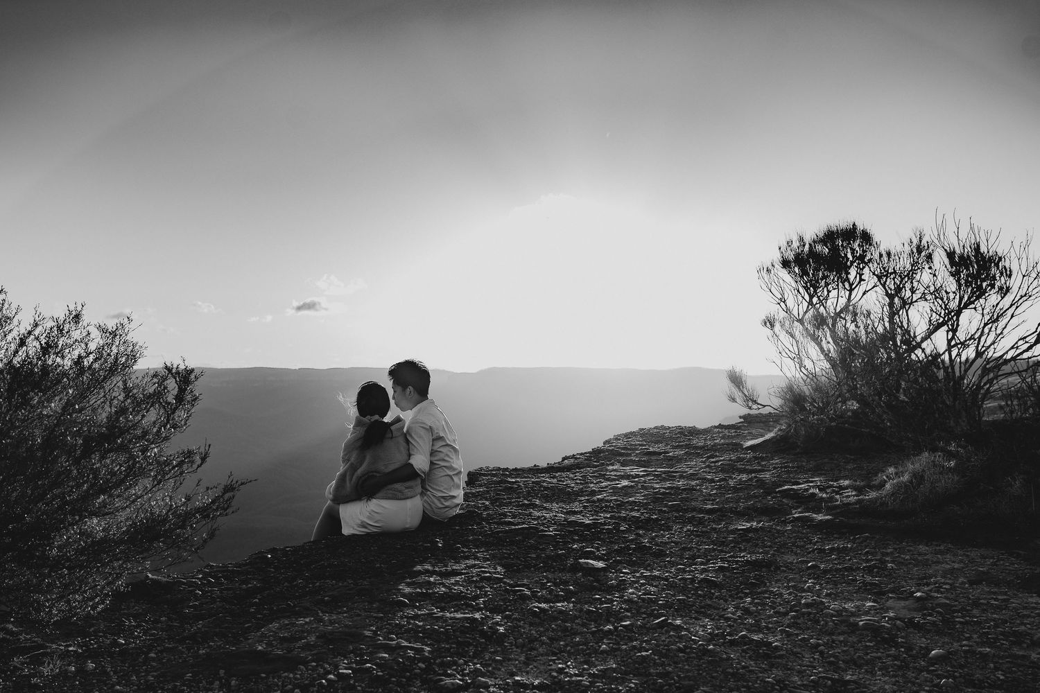 A silhouetted pair sits close together on a clifftop overlooking a vast mountain landscape.