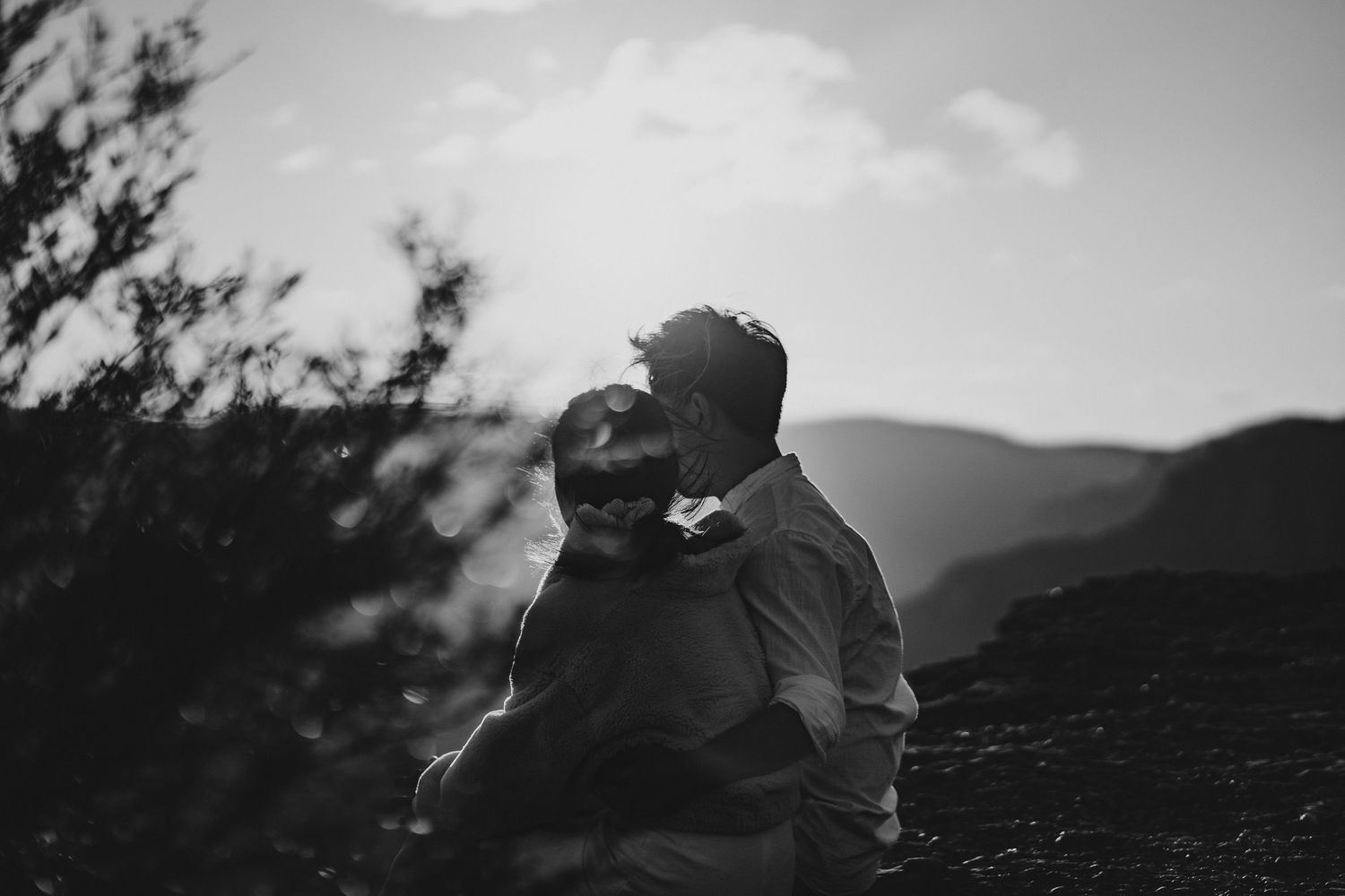 A romantic black and white portrait of a couple embracing against a mountain backdrop.