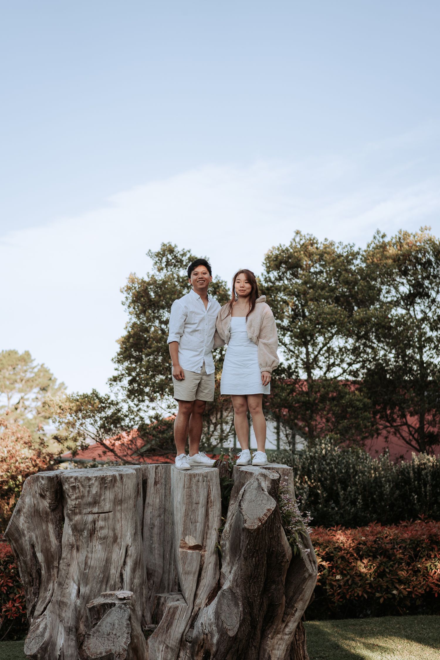 A couple in matching white outfits stands together on a large tree stump outdoors.