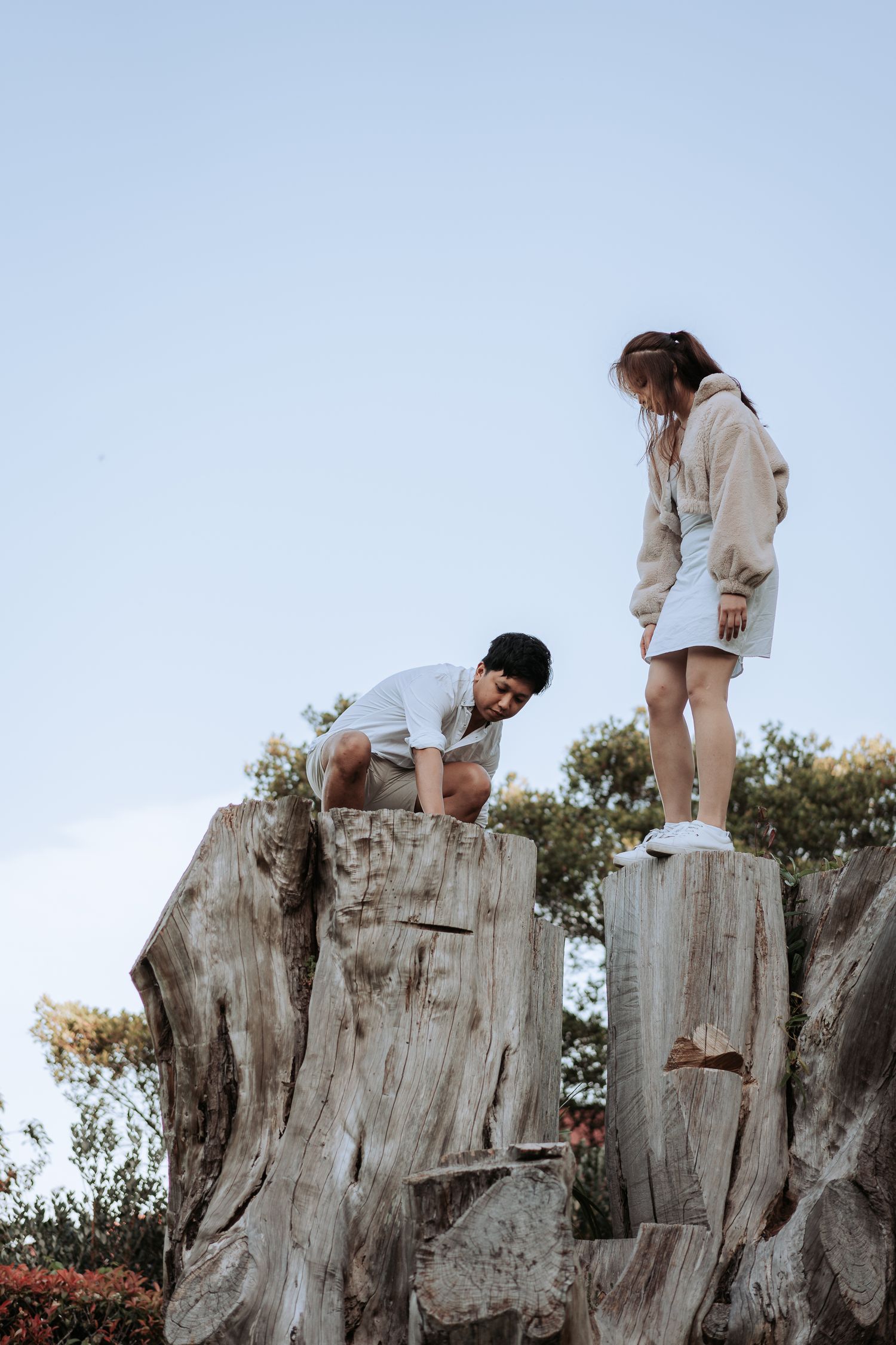 Two people in white clothing climb on a large weathered tree stump against a blue sky.
