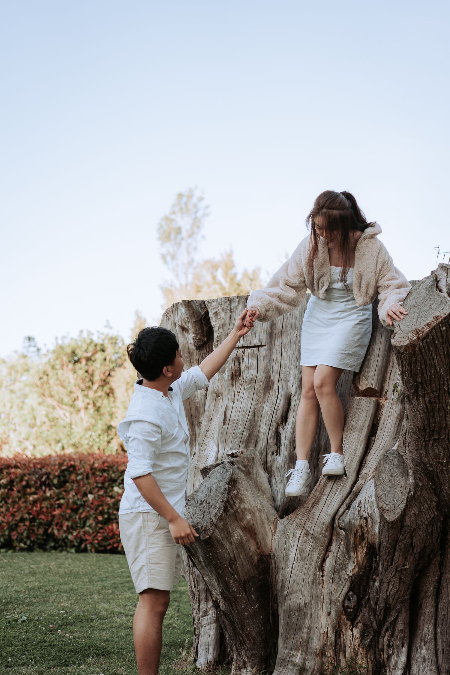 A couple helps each other navigate a large tree stump in a garden setting.