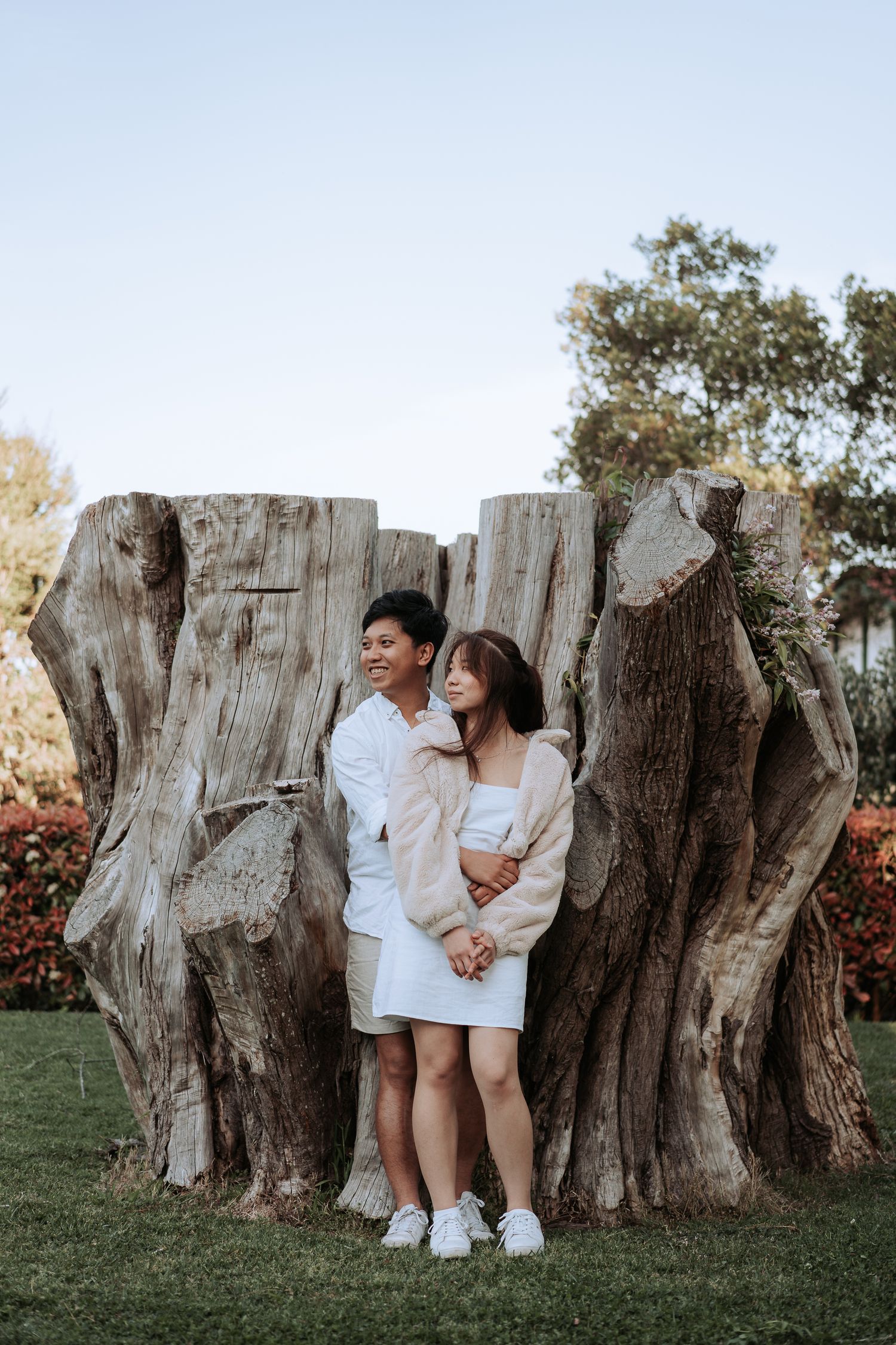 A couple in white outfits poses together against a large tree stump in a garden.