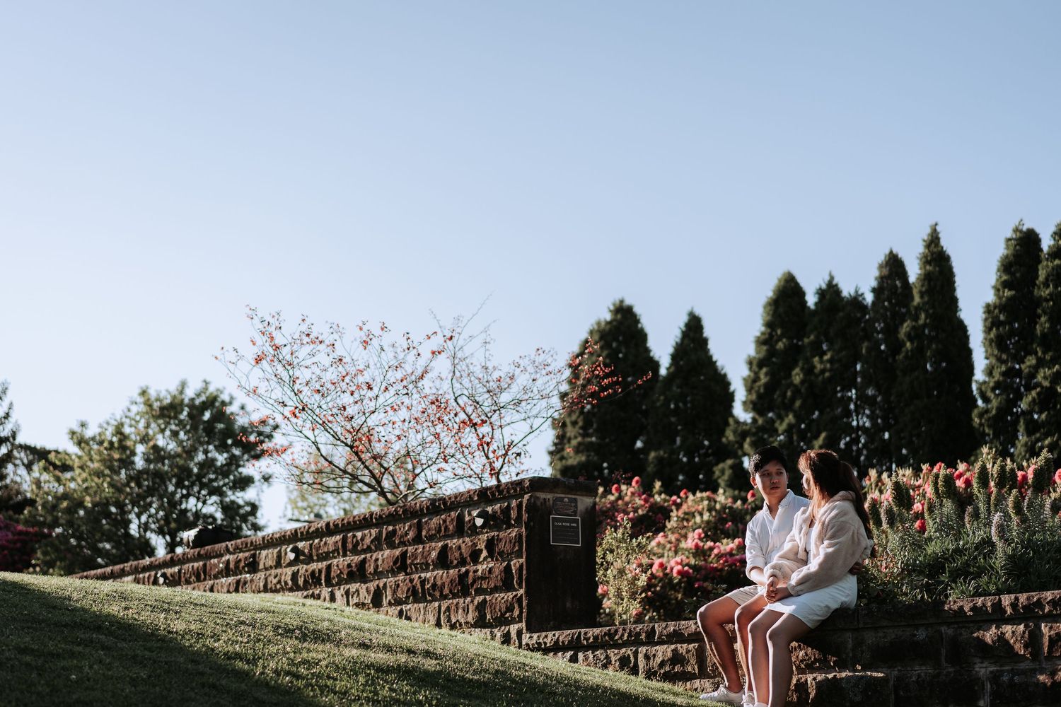 Two people in white clothing sitting on stone steps in a garden with evergreen trees.