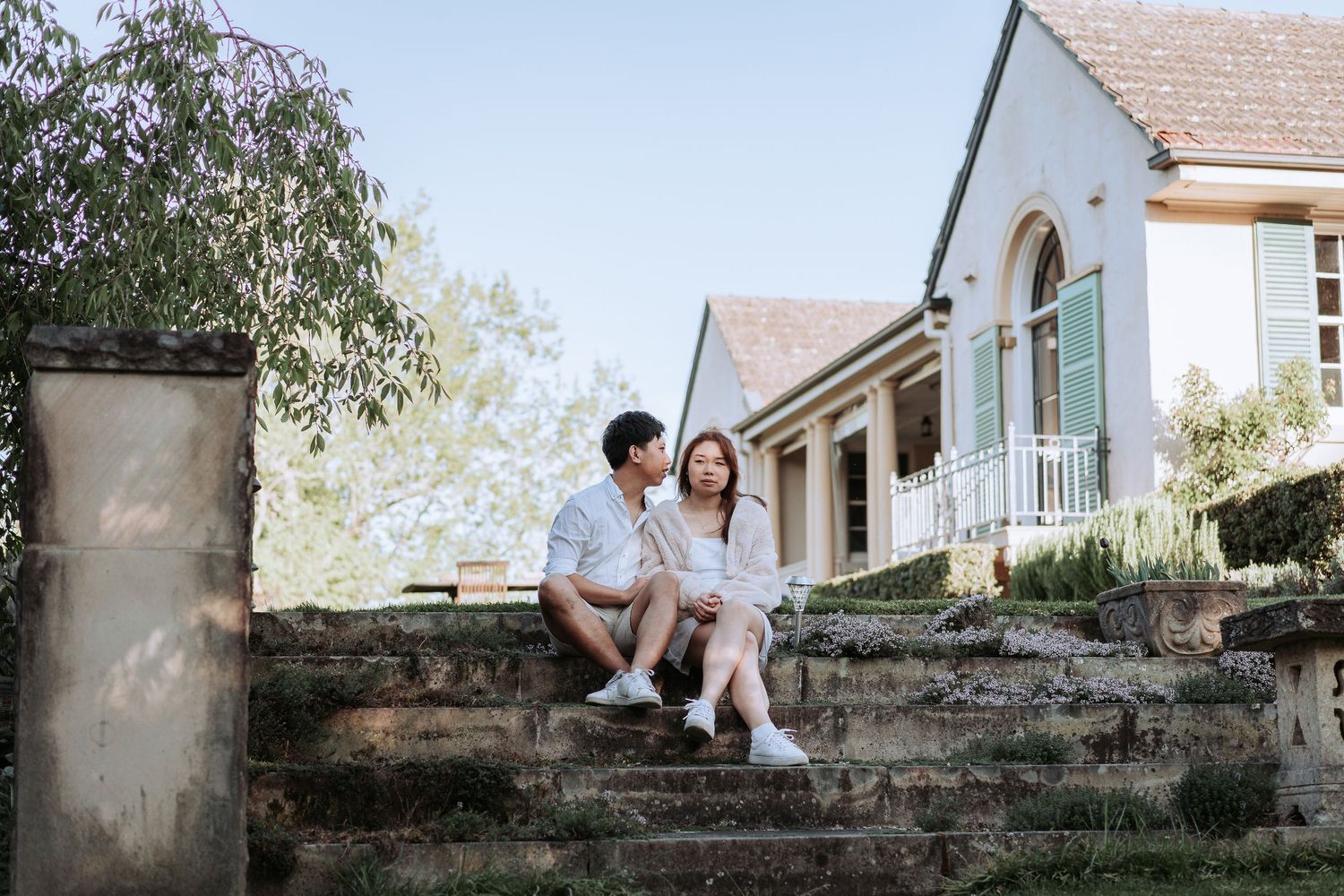 A couple in white clothes sits together on stone steps leading to a charming cottage.