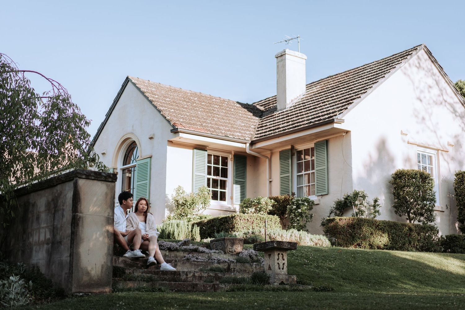 A couple relaxes on stone steps outside a quaint white cottage with blue trim.