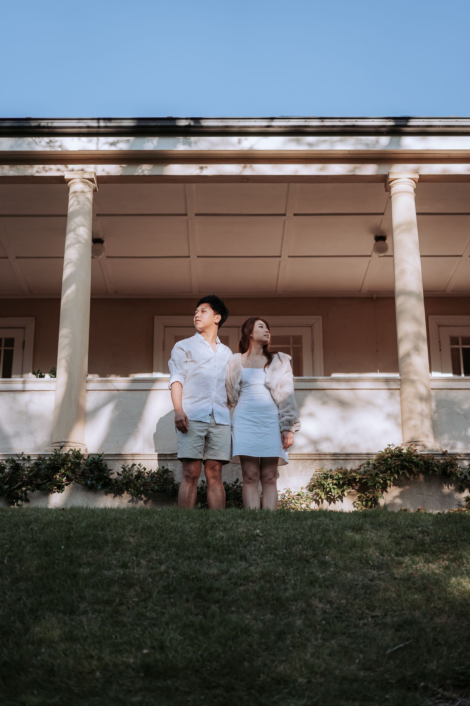 A couple stands together on a lawn in front of a classical columned porch.