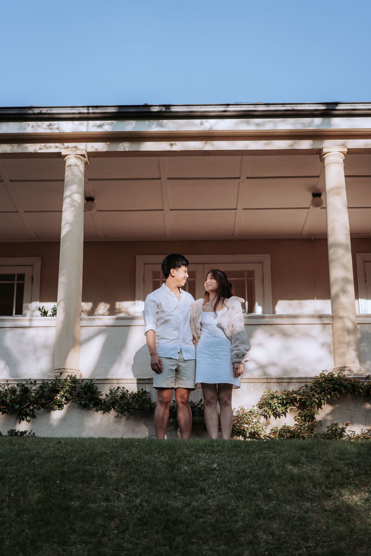 A couple in coordinated outfits poses in front of a columned porch during golden hour.