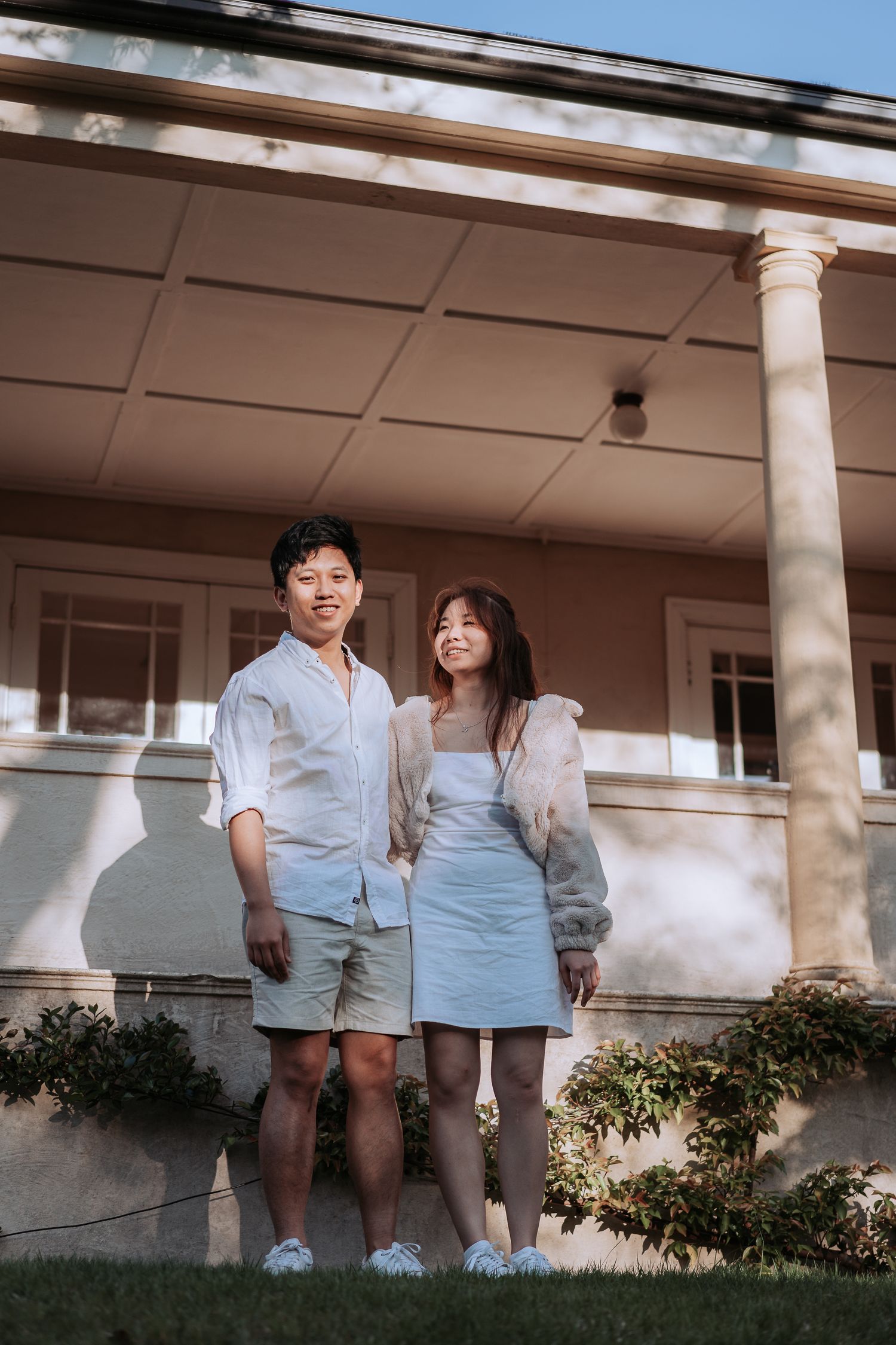 A couple in matching light-colored outfits stands together on a lawn near white columns.