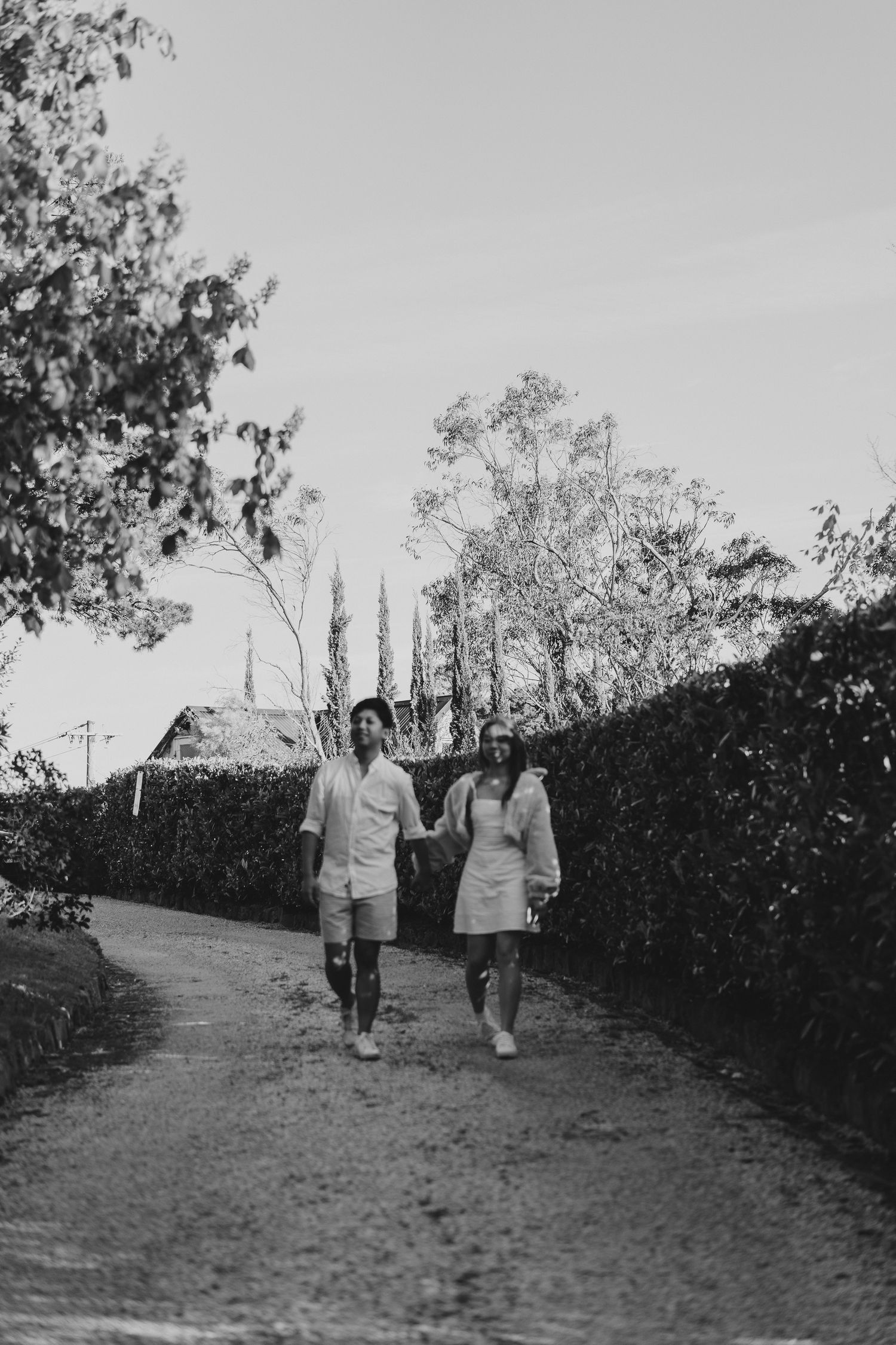 A black and white photo of a couple walking together on a garden path lined with hedges.