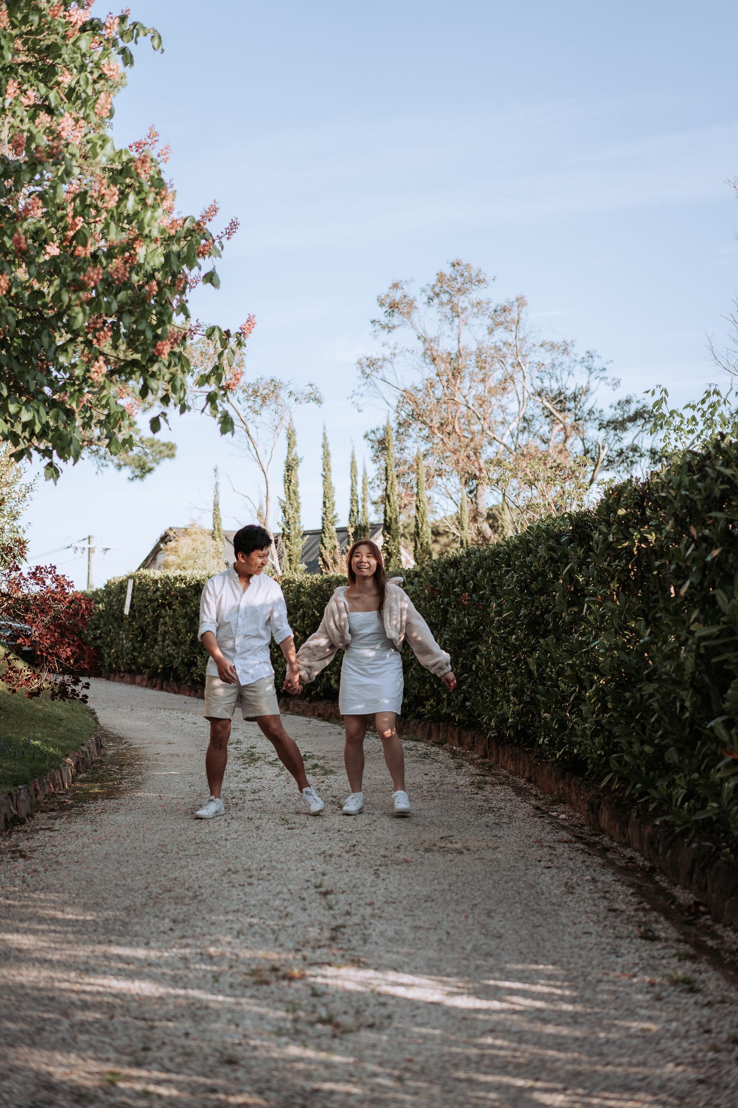 A playful couple in matching white outfits walking along a winding garden path with tall hedges.